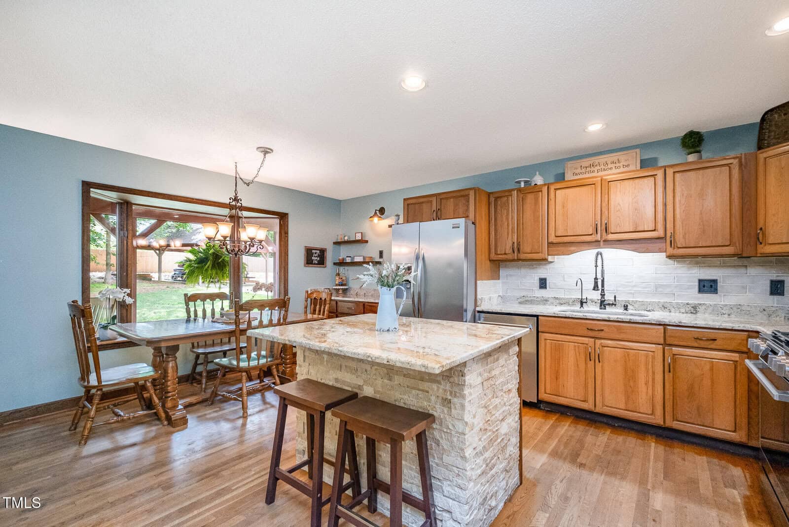 Bright kitchen with wooden cabinets, stainless steel appliances, an island with a stone base and two stools, and a dining area with a wooden table and chairs by a large window overlooking a green yard.