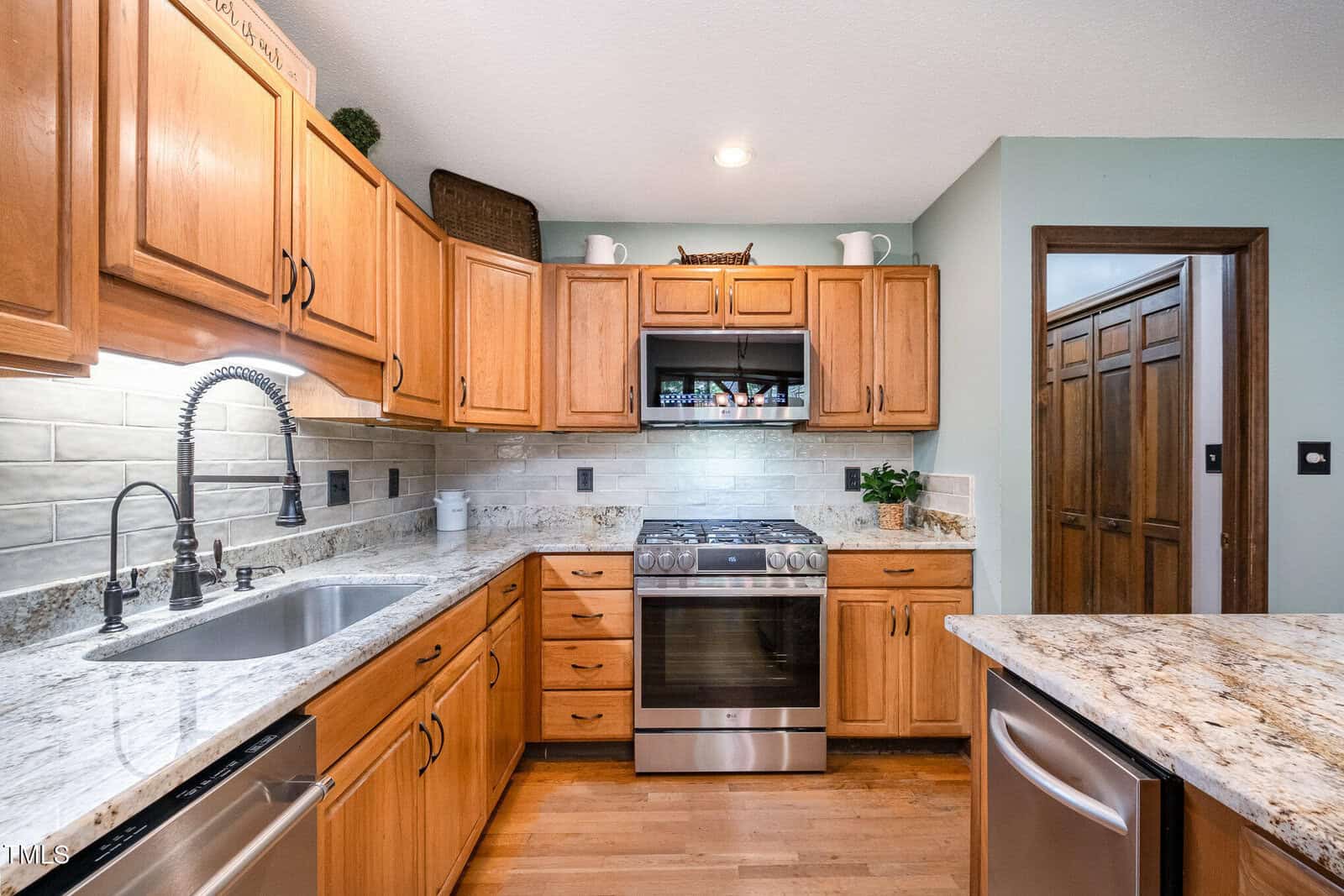 Modern kitchen with wooden cabinets, stainless steel appliances, granite countertops, tile backsplash, a double sink, and a gas stove. The walls are light green with wood floors and some decorative items on top of the cabinets.
