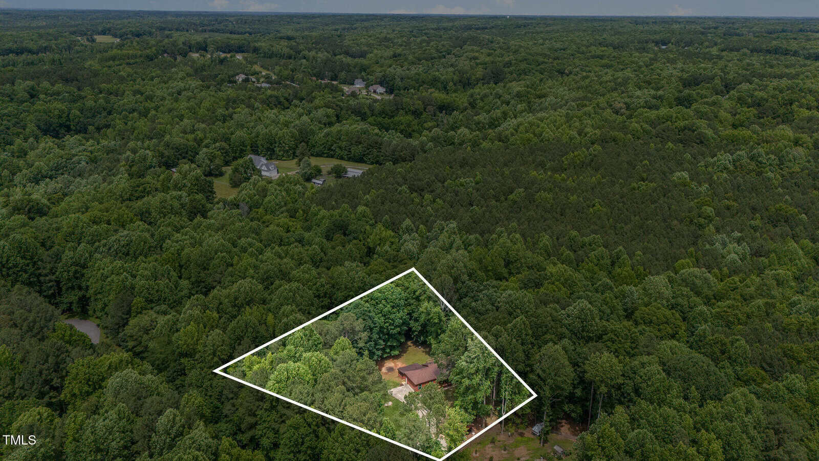 Aerial view of a densely wooded area with a house and property outlined in white near the bottom center, surrounded by trees and several other houses visible in the distance.