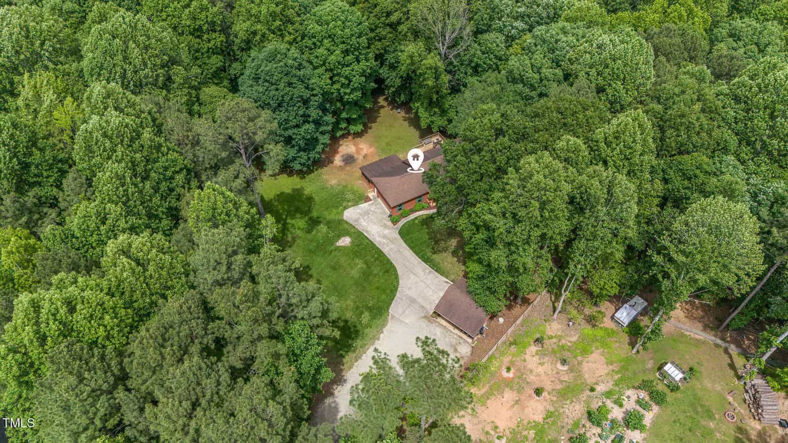 Aerial view of a house surrounded by dense green trees, with a curved driveway leading to the garage and a cleared yard area around the home. Some outdoor structures are visible among the trees.