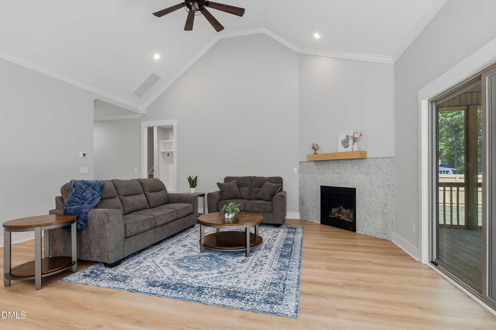 Modern living room with gray sofas, a blue patterned rug, round wooden tables, a fireplace with a tile surround, light wood floors, vaulted ceiling, ceiling fan, and large windowed doors opening to a screened porch.