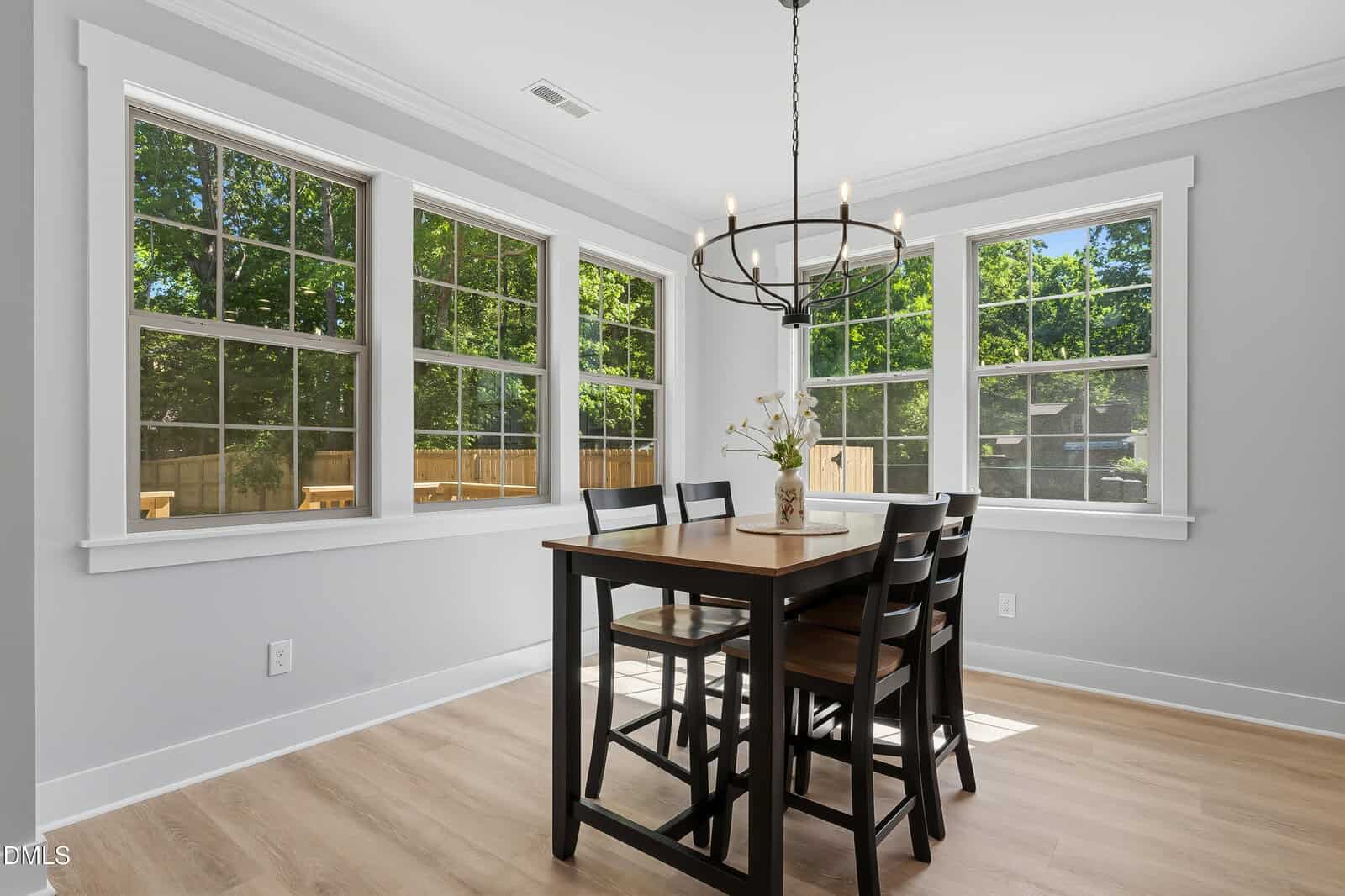 Bright dining room with large windows, black chandelier, wooden table with four black chairs, and a small vase with flowers; light wood floor and white trim, with leafy trees visible outside.