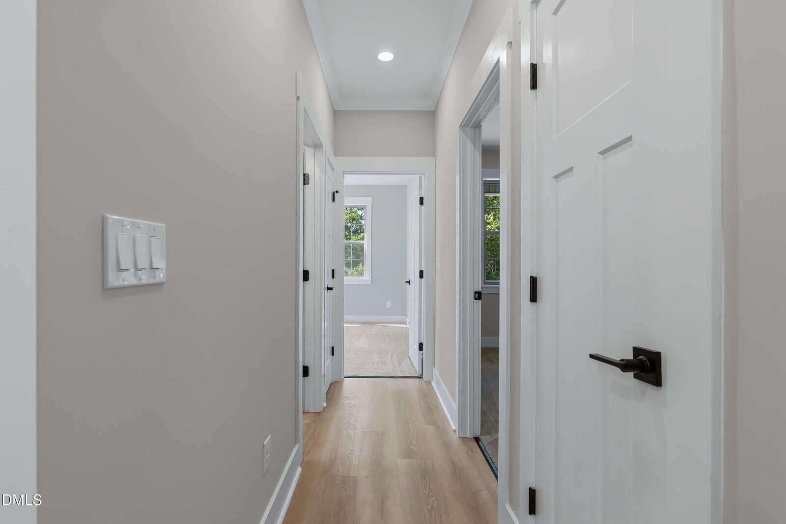 A hallway with light wood floors, neutral walls, white doors on both sides, and a view into bright rooms at the end. A triple light switch is on the left wall. Natural light comes in from windows ahead.