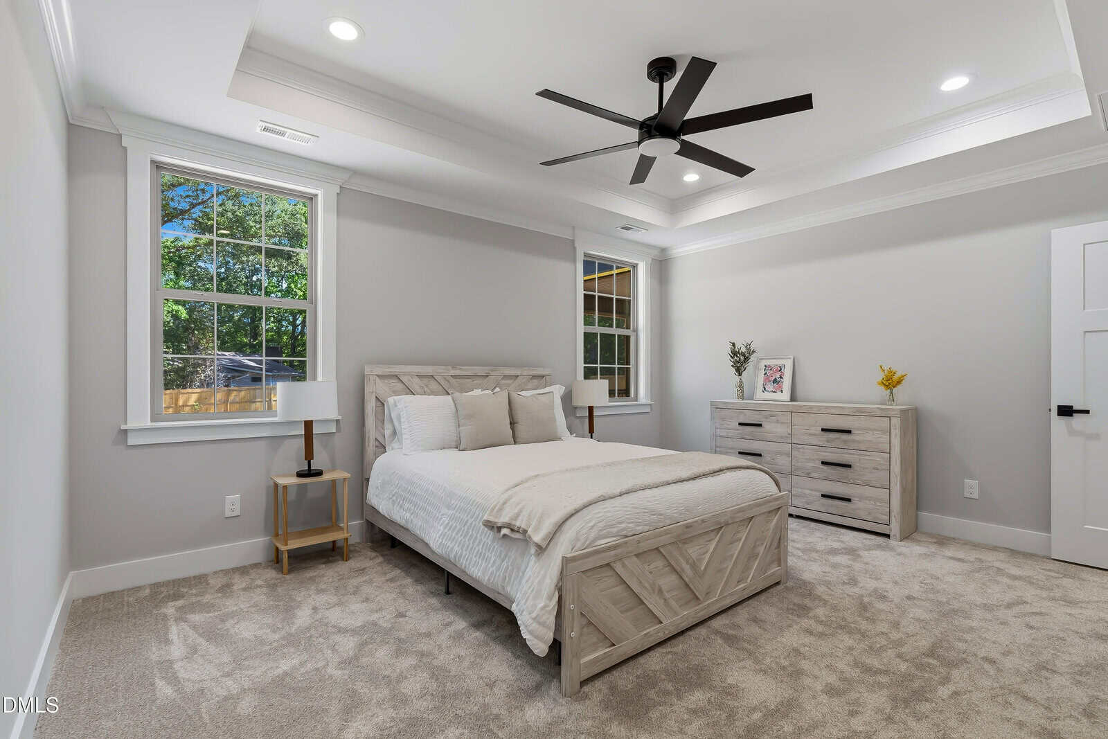 Bright bedroom with a queen bed, matching wooden nightstand and dresser, beige carpet, a ceiling fan, and two large windows letting in natural light. Decor includes a plant, books, and a small tray on the dresser.
