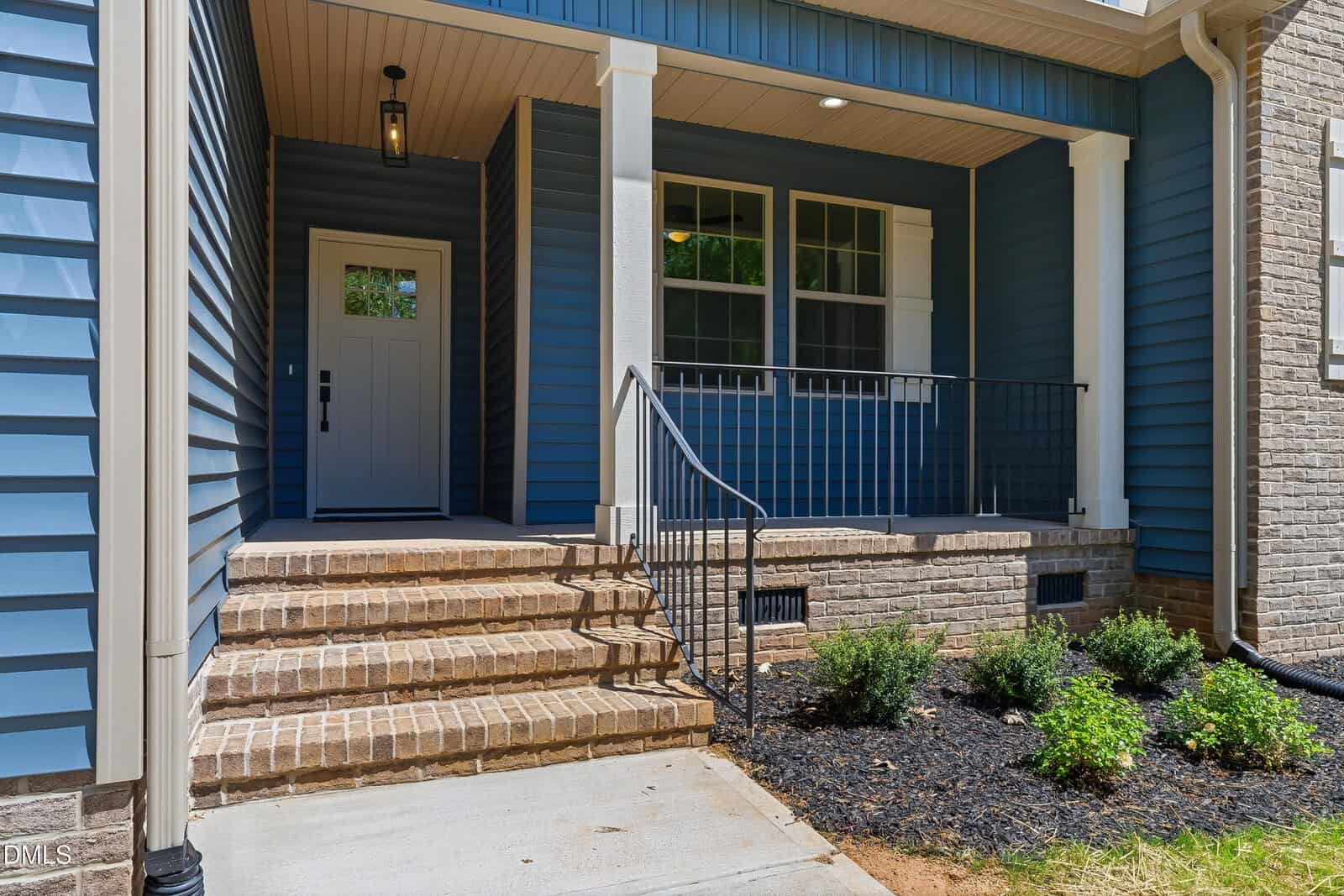 Front porch of a house with blue siding, white trim, brick steps and foundation, black metal railing, a white front door with sidelights, and a small landscaped area with shrubs.