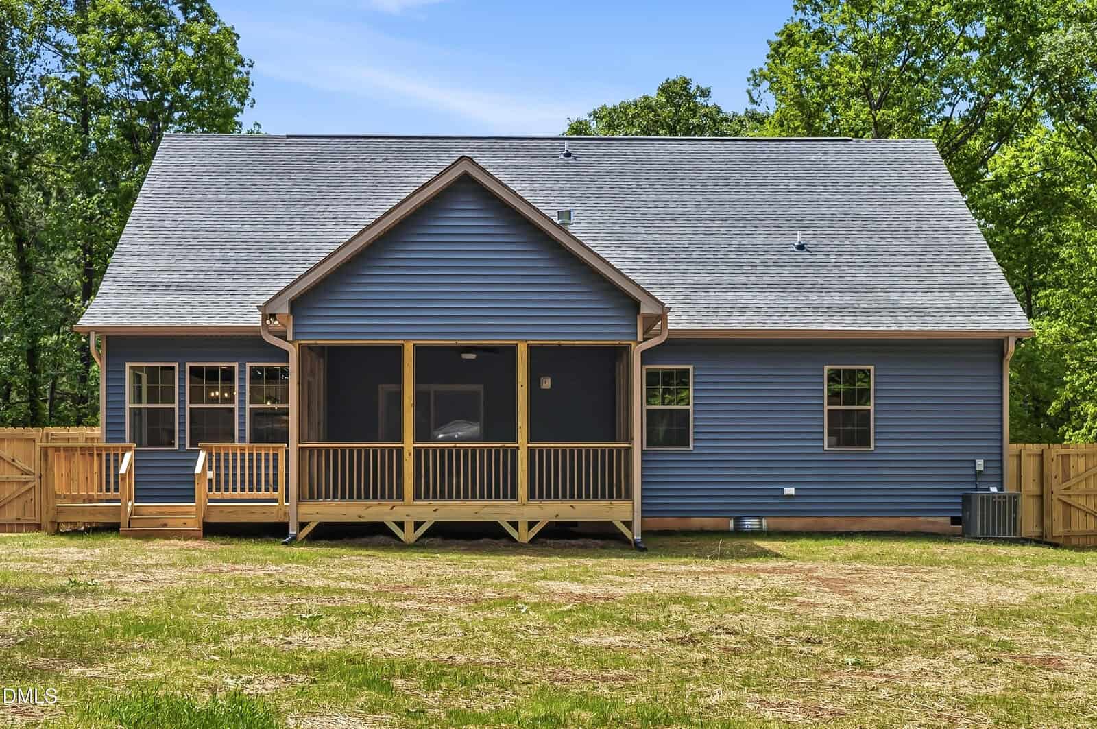 A single-story blue house with a gray roof, wooden screened-in back porch, fenced yard, and surrounded by green trees under a clear sky.