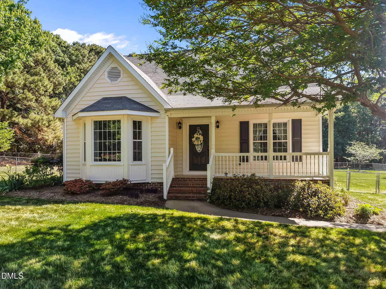 A small, single-story yellow house with white trim, a bay window, a front porch with a wreath on the door, brick steps, and a well-kept lawn, shaded by a large tree, on a sunny day.