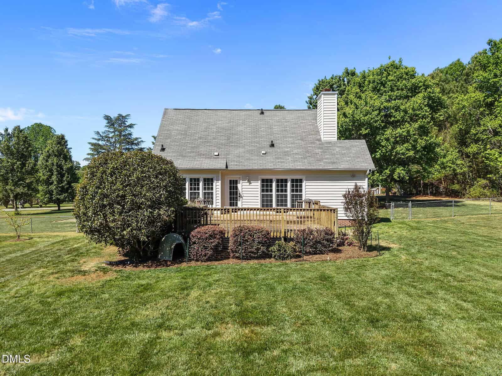 White house with gray roof, chimney, and a wooden deck sits in a large, well-maintained grassy yard with trees and bushes on a sunny day. Fence and more trees are visible in the background.