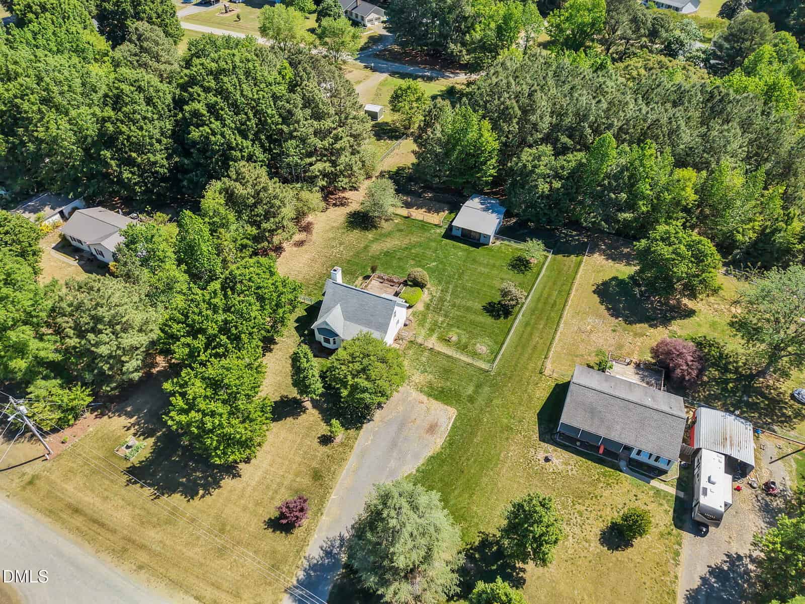 Aerial view of a rural residential area with three houses surrounded by trees, large green lawns, a driveway, and a shed. The landscape is lush and well-maintained under bright daylight.