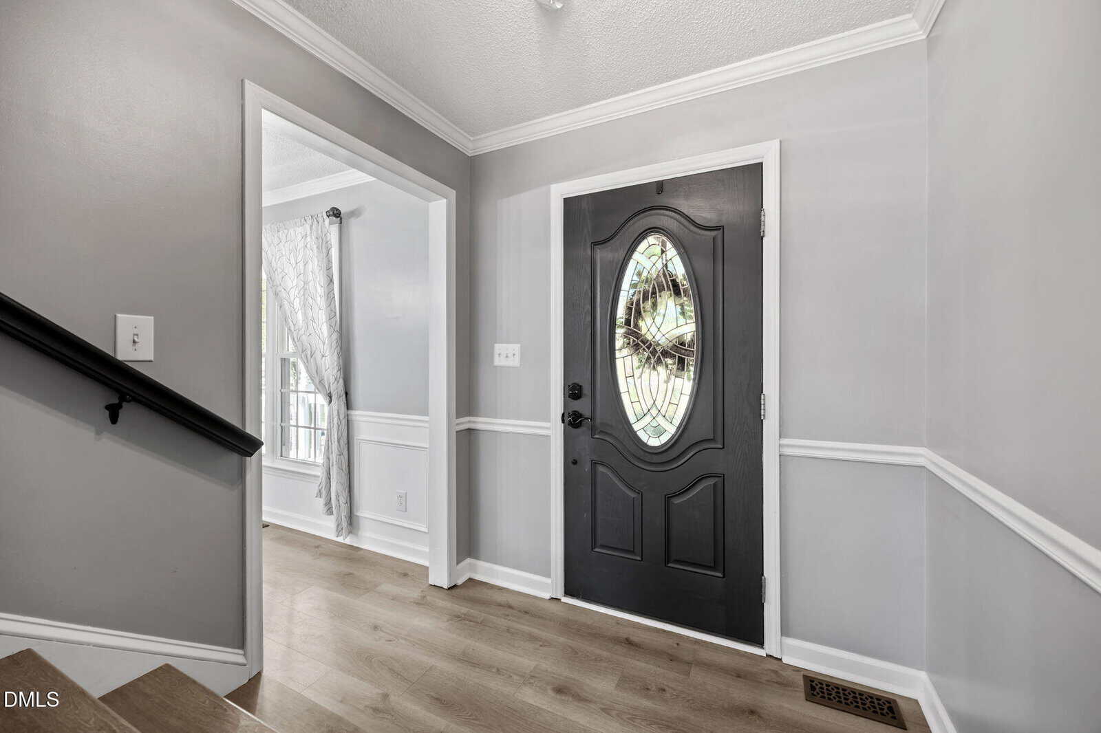Entryway with light wood flooring, gray walls, white trim, and a black front door with an oval stained glass window. Stairs are to the left and a sunlit room with curtains is visible through an open doorway.