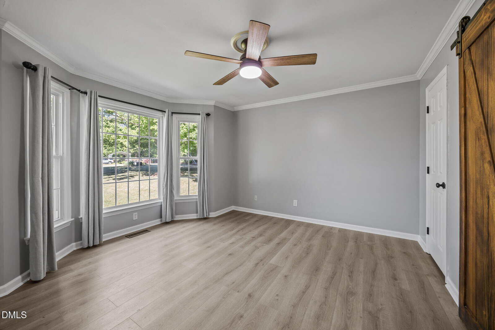 Bright, empty room with light wood floors, gray walls, a ceiling fan, bay window with gray curtains, white trim, and a wooden sliding barn door. Natural light fills the space, creating an airy and modern feel.