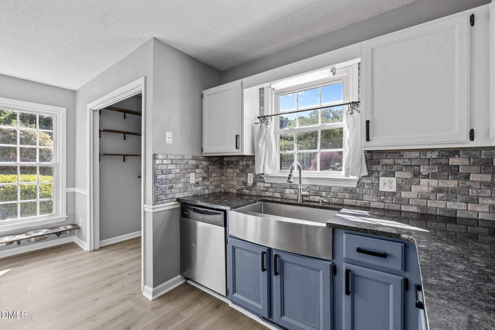 Bright kitchen with a farmhouse sink, blue lower cabinets, white upper cabinets, stainless dishwasher, gray stone backsplash, and a large window with a view outside; adjacent pantry with shelves visible.