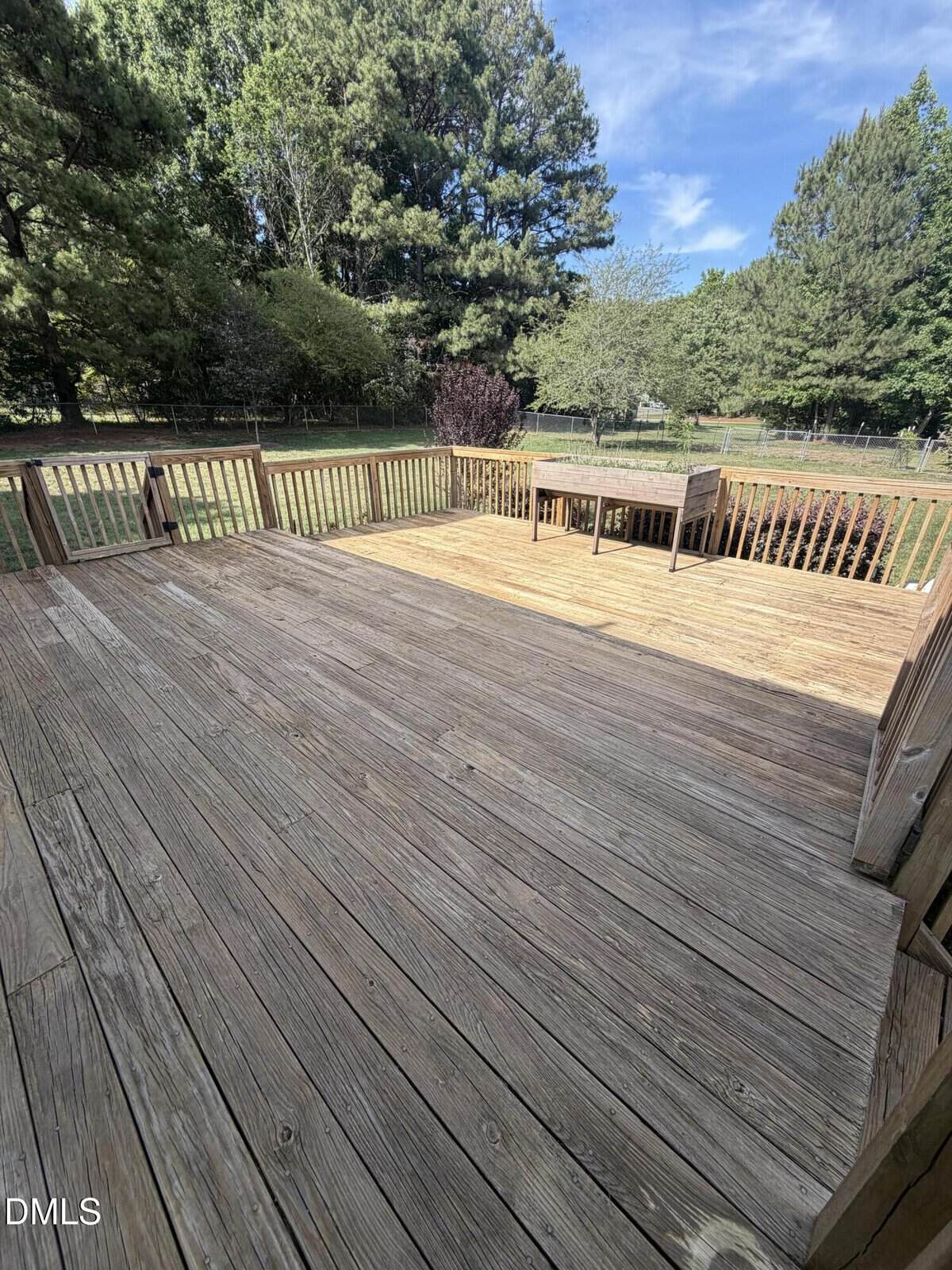 Wooden backyard deck with railing, overlooking a grassy yard surrounded by trees. A wooden table and benches are at the far end of the deck under a clear, sunny sky.