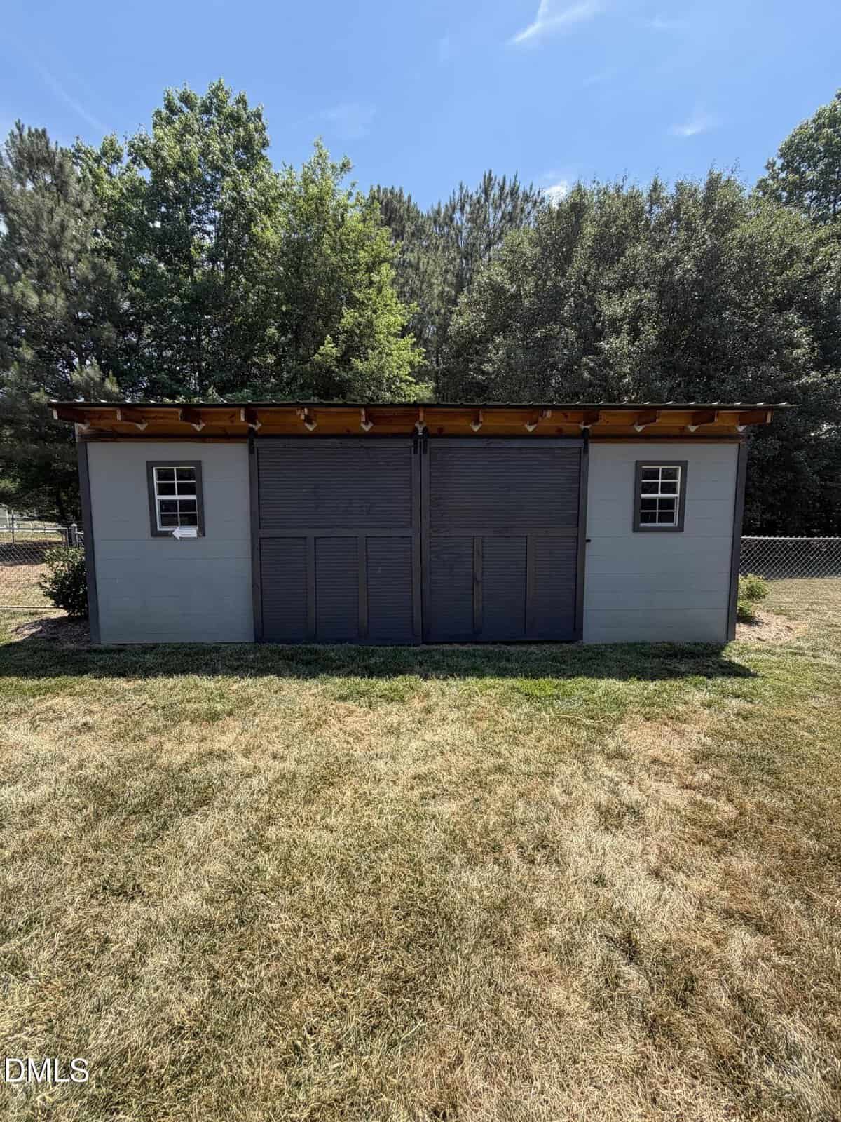A light gray shed with two central dark double doors and two small windows, one on each side. The shed has a wooden roof and sits on a grassy lawn, with trees and a chain-link fence in the background.