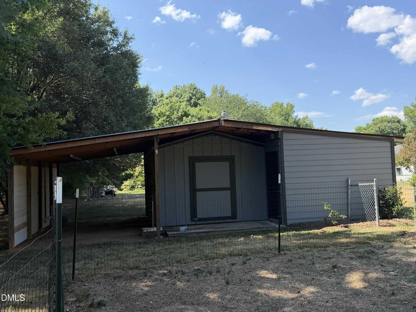 A small gray barn or shed with a slanted roof sits in a fenced yard, surrounded by trees and grass under a blue sky with scattered clouds.