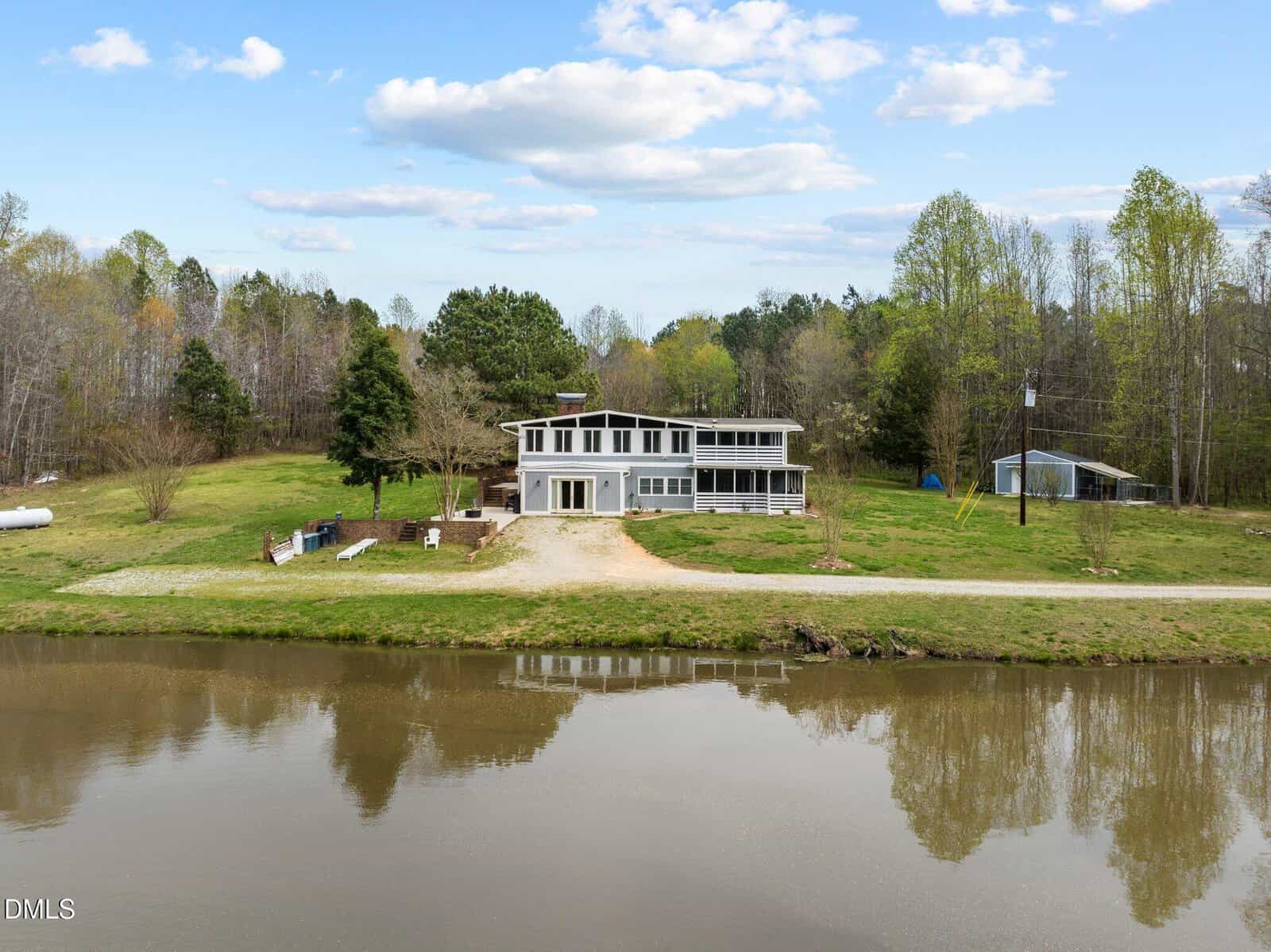 A modern white house with large windows and a screened porch sits beside a calm pond, surrounded by green trees and grass under a partly cloudy sky. A gravel driveway leads to the house.