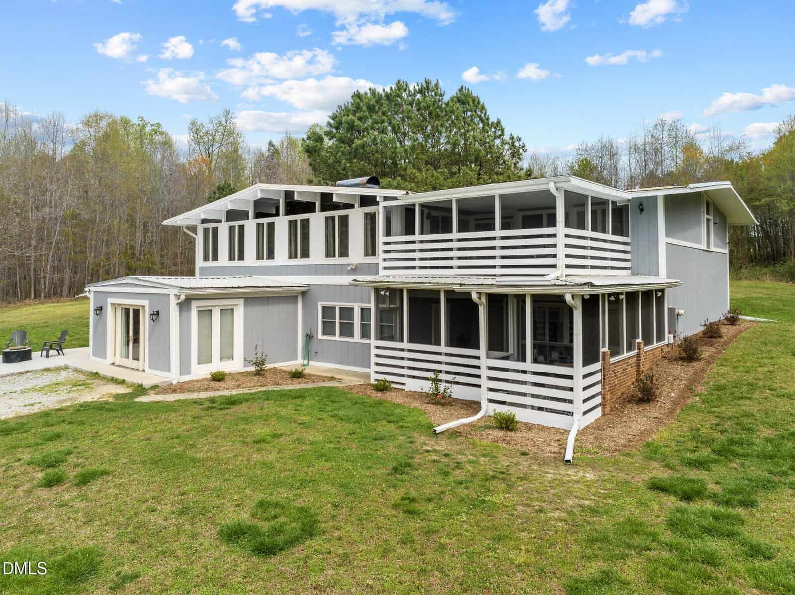 Two-story gray house with white trim and large windows, featuring two covered balconies and a patio area. The home sits on a spacious grassy lawn surrounded by trees under a partly cloudy sky.