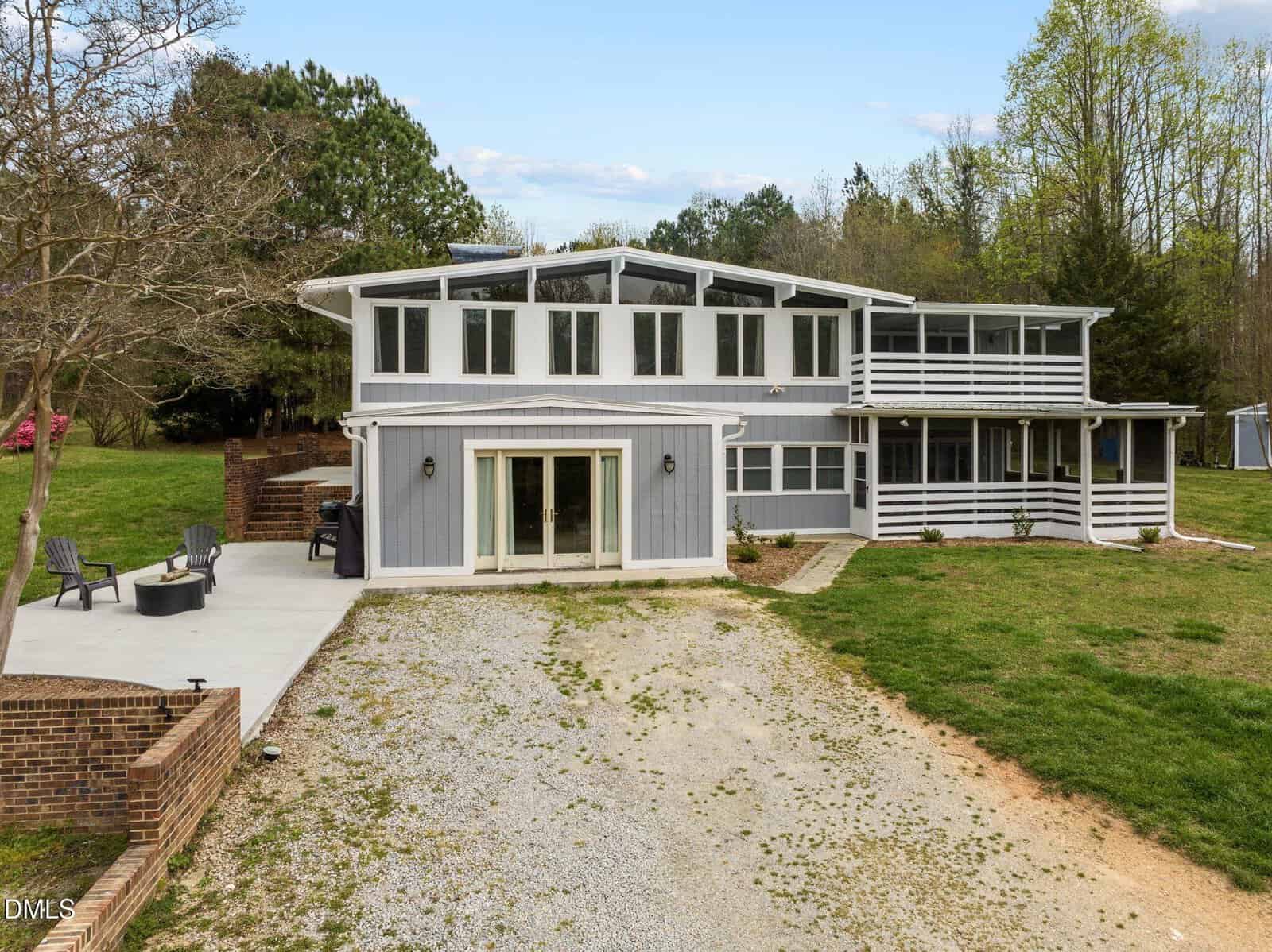 A modern two-story gray house with large windows, a stone patio with seating, a gravel driveway, and a screened porch, surrounded by grass, trees, and a partially cloudy sky.