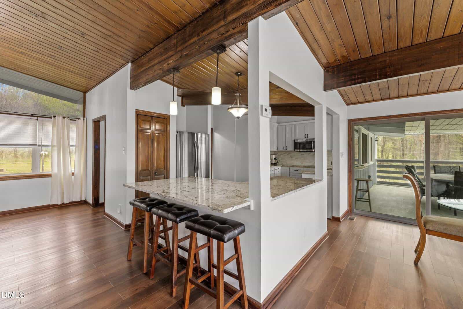 Modern kitchen with wood floors and ceiling, white walls, granite island with three stools, stainless steel appliances, and open view to dining and sunroom areas. Large windows bring in natural light.