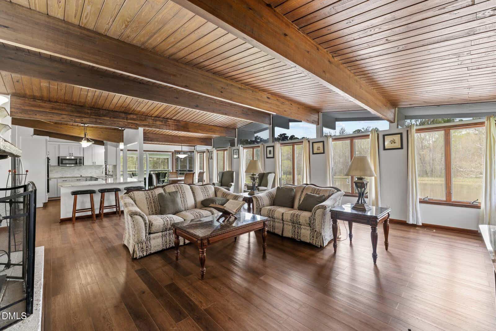 Spacious living room with wood floors and ceiling, exposed beams, large windows, and cozy patterned sofas with wooden coffee and side tables. Kitchen visible in the background. Bright, natural light fills the space.
