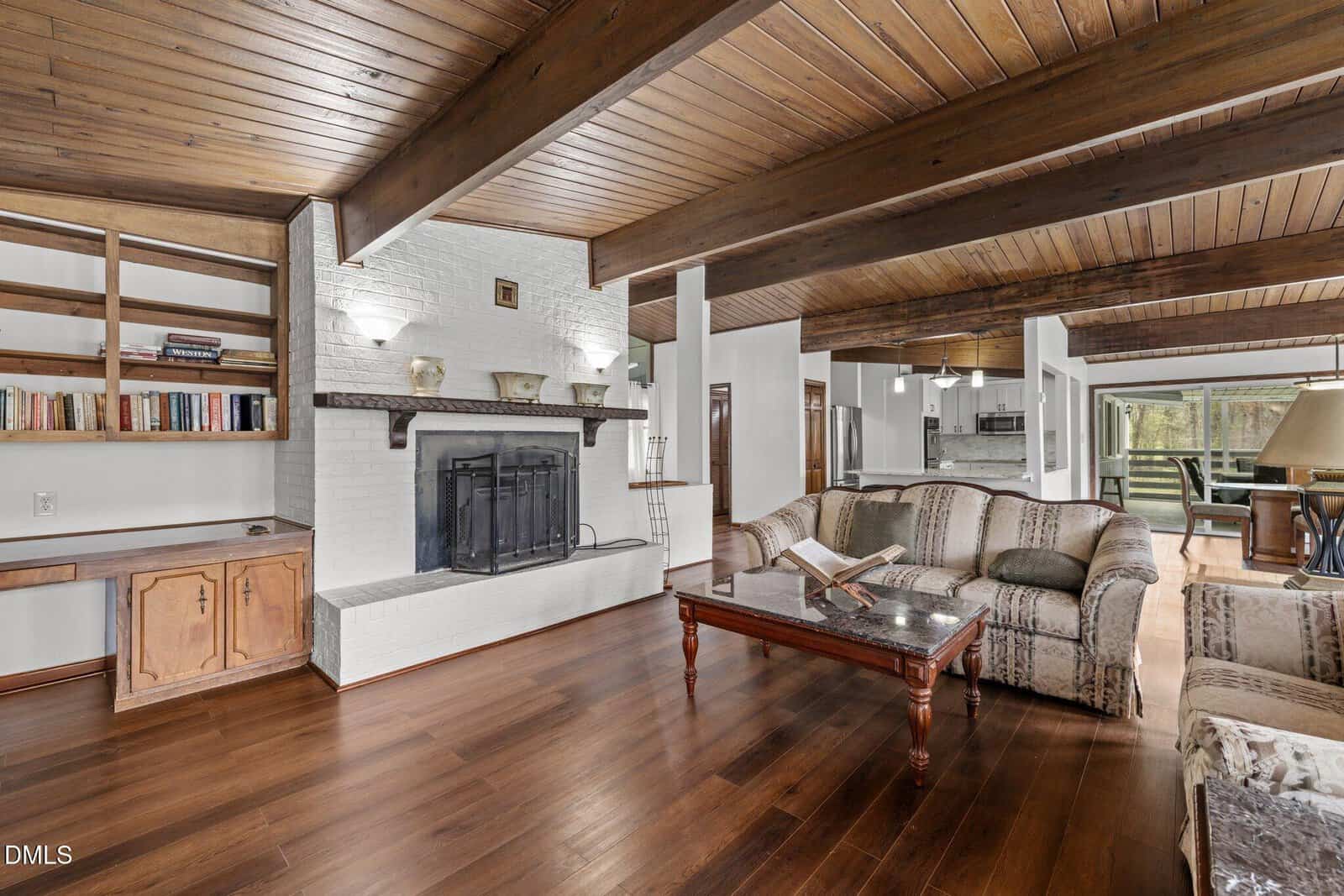 A cozy living room with wood floors, exposed wood ceiling beams, a white brick fireplace, built-in shelves with books, patterned sofas, a coffee table, and a view into the dining area and kitchen.