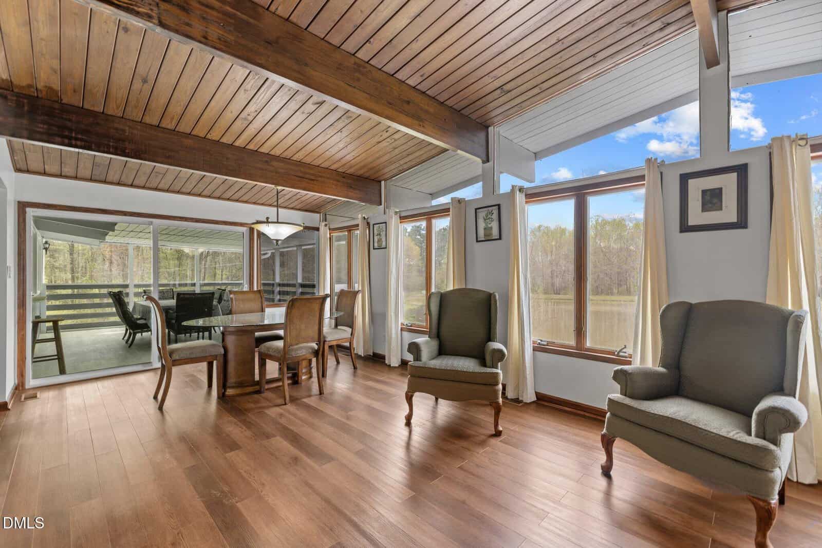 A bright dining room with wood floors, exposed beams, large windows with lake views, a dining table with six chairs, and two armchairs near pale curtains. A sunroom with more seating is visible through glass doors.