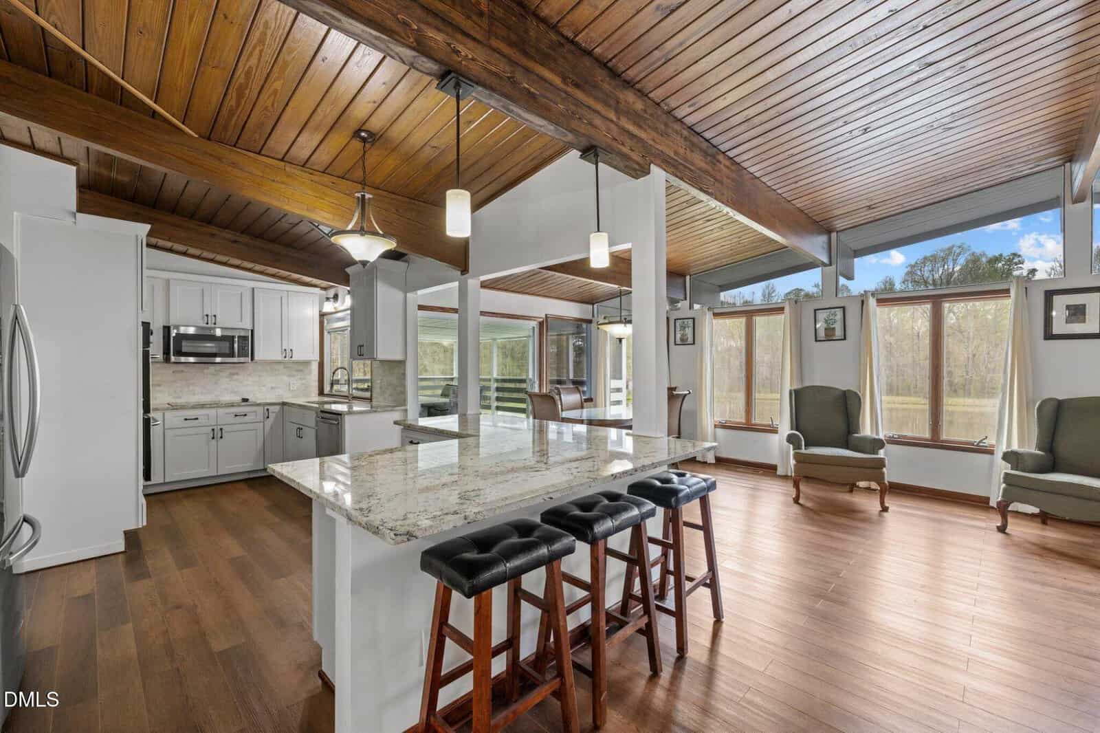 Spacious kitchen with white cabinets, granite island, and pendant lights, open to a living area with large windows, wood ceilings, and hardwood floors. Three stools line the island; two armchairs sit by the windows.