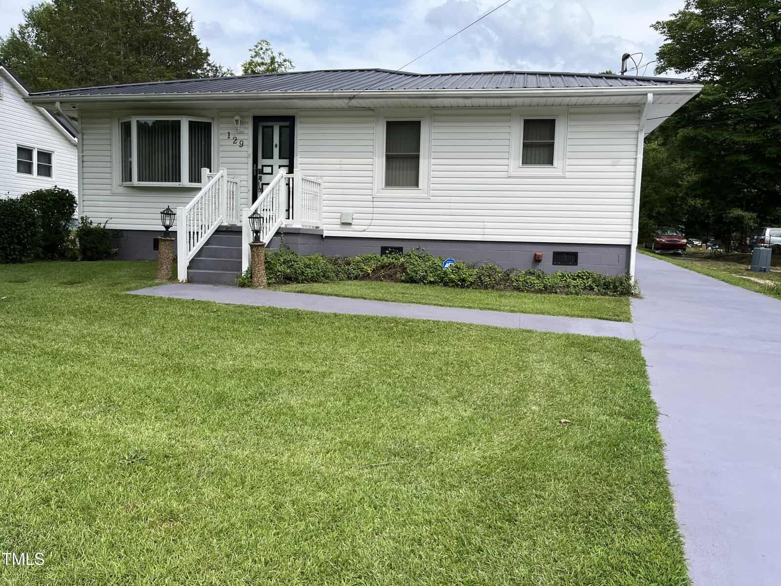 A single-story white house with a dark roof, steps leading to the front door, and a well-maintained green lawn. A paved driveway runs along the right side of the house. Shrubs border the front of the house.