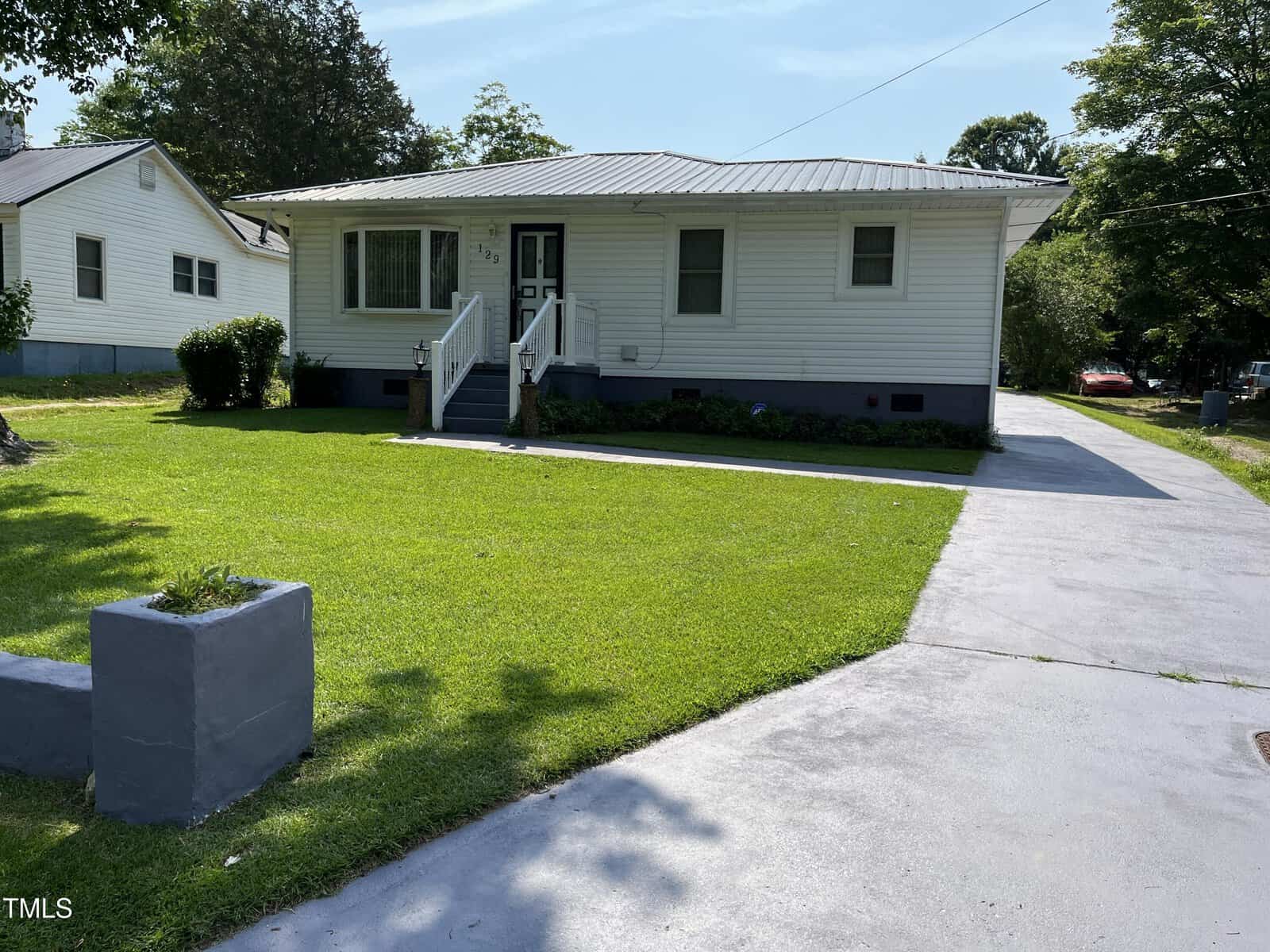 Single-story white house with a metal roof, a small front porch with steps and railings, and a wide concrete driveway leading to the street. The lawn is neatly mowed and there are a few shrubs along the house.
