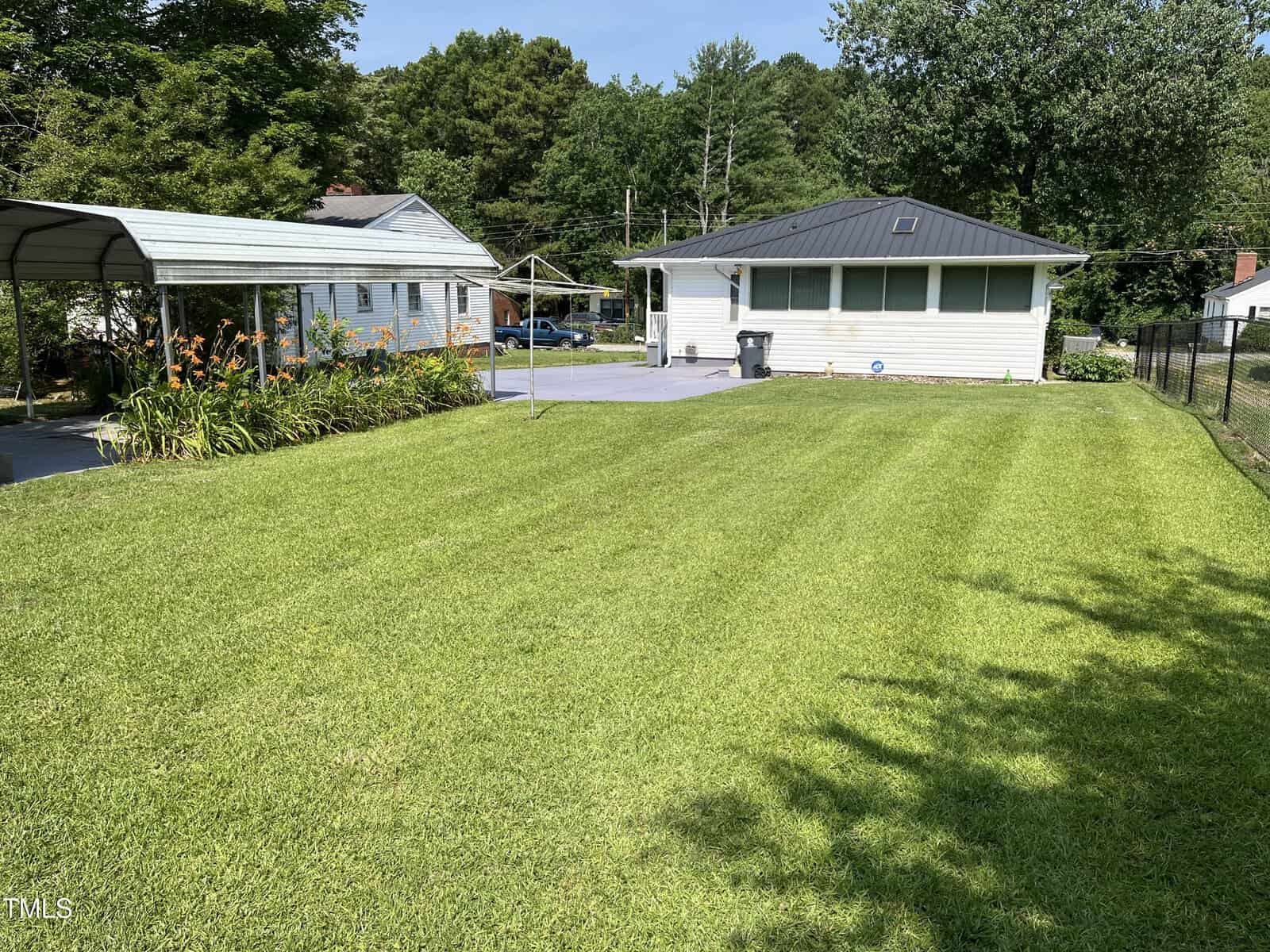 A neatly mowed green lawn stretches toward a single-story white house with a black roof, a paved driveway, a metal carport, and a clothesline, surrounded by trees and a wire fence on a sunny day.