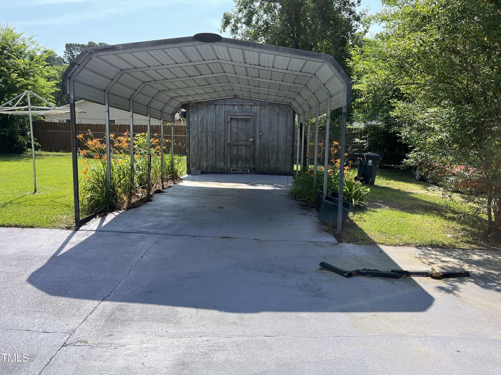 A metal carport with a curved roof stands over a concrete driveway, leading to a small wooden shed. Orange flowers line both sides of the carport, and green grass and trees surround the area.