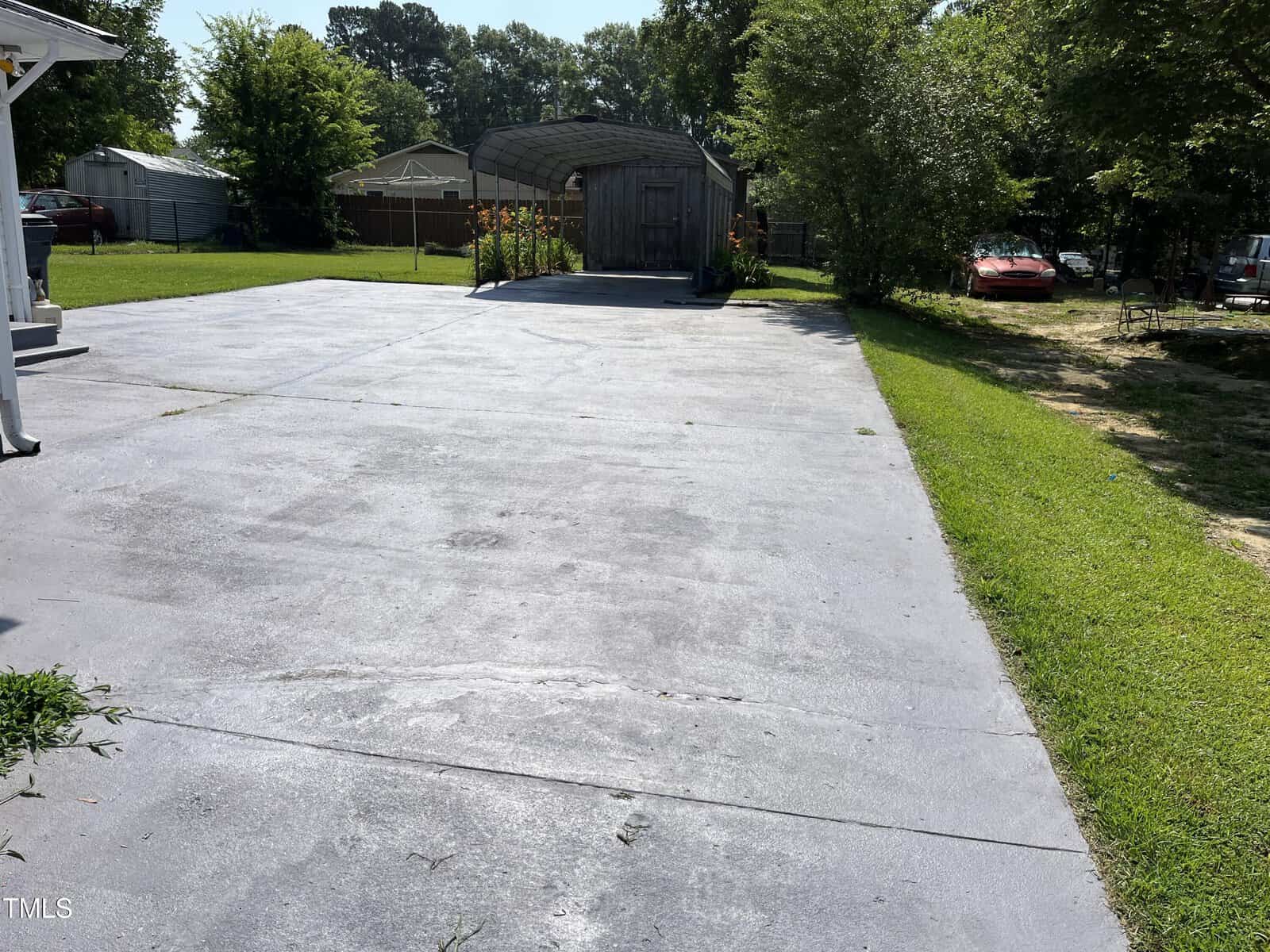 A wide concrete driveway leads to a metal carport and wooden shed in a backyard, with green grass and trees on both sides. A red car is partially visible in the background.