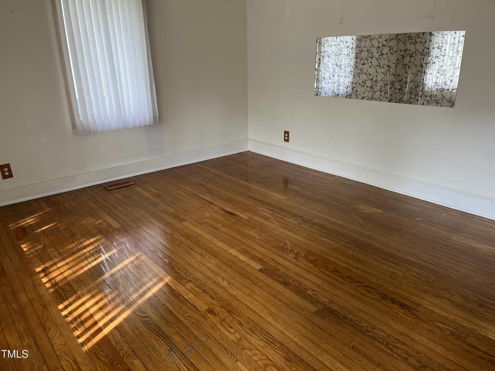 Empty room with hardwood floors, white walls, a window with blinds letting in sunlight, and a wall mirror reflecting patterned curtains.