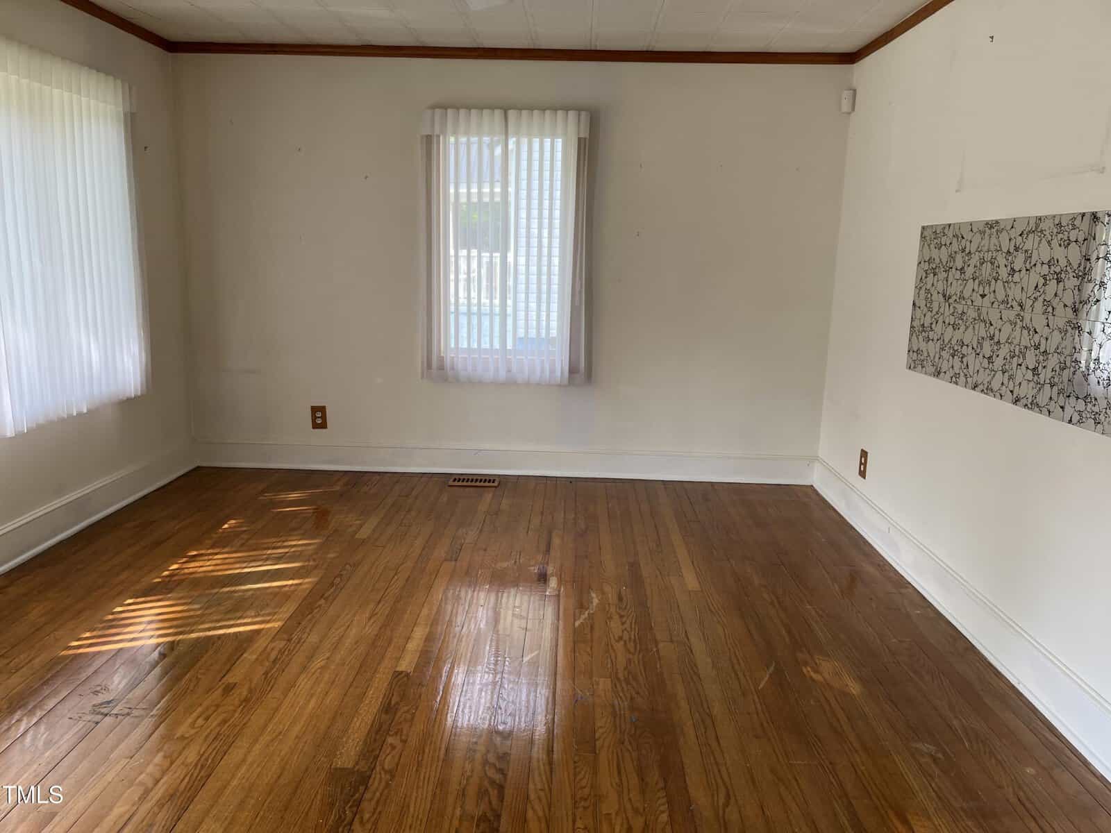 Empty room with hardwood floors, two windows with vertical blinds letting in sunlight, and a patterned panel on the right wall. Walls and trim are white, and there is a floor vent near one window.