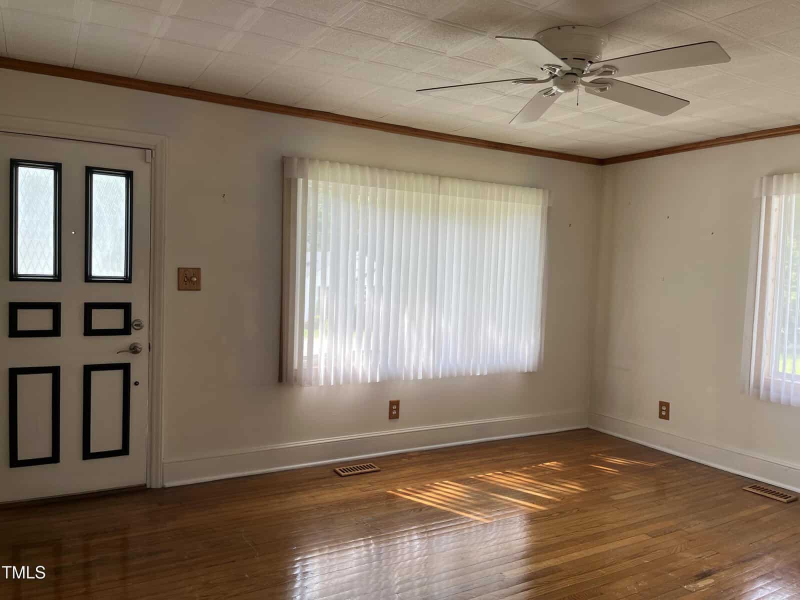 Empty room with hardwood floors, white walls, a ceiling fan, a window with vertical blinds, and a door with glass panels. Sunlight shines through the window, creating bright patches on the floor.