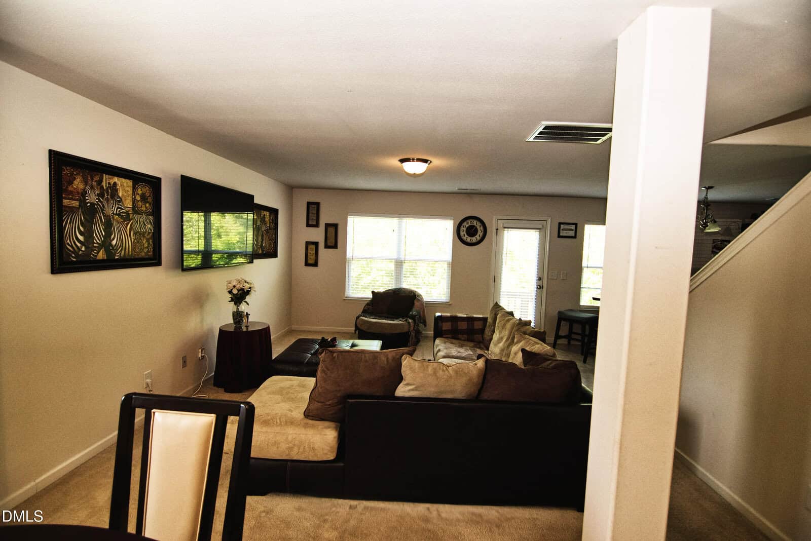 A spacious living room with a sectional sofa, wall art, a flat-screen TV, large windows letting in natural light, and a door leading outside. Neutral tones and contemporary decor create a cozy atmosphere.
