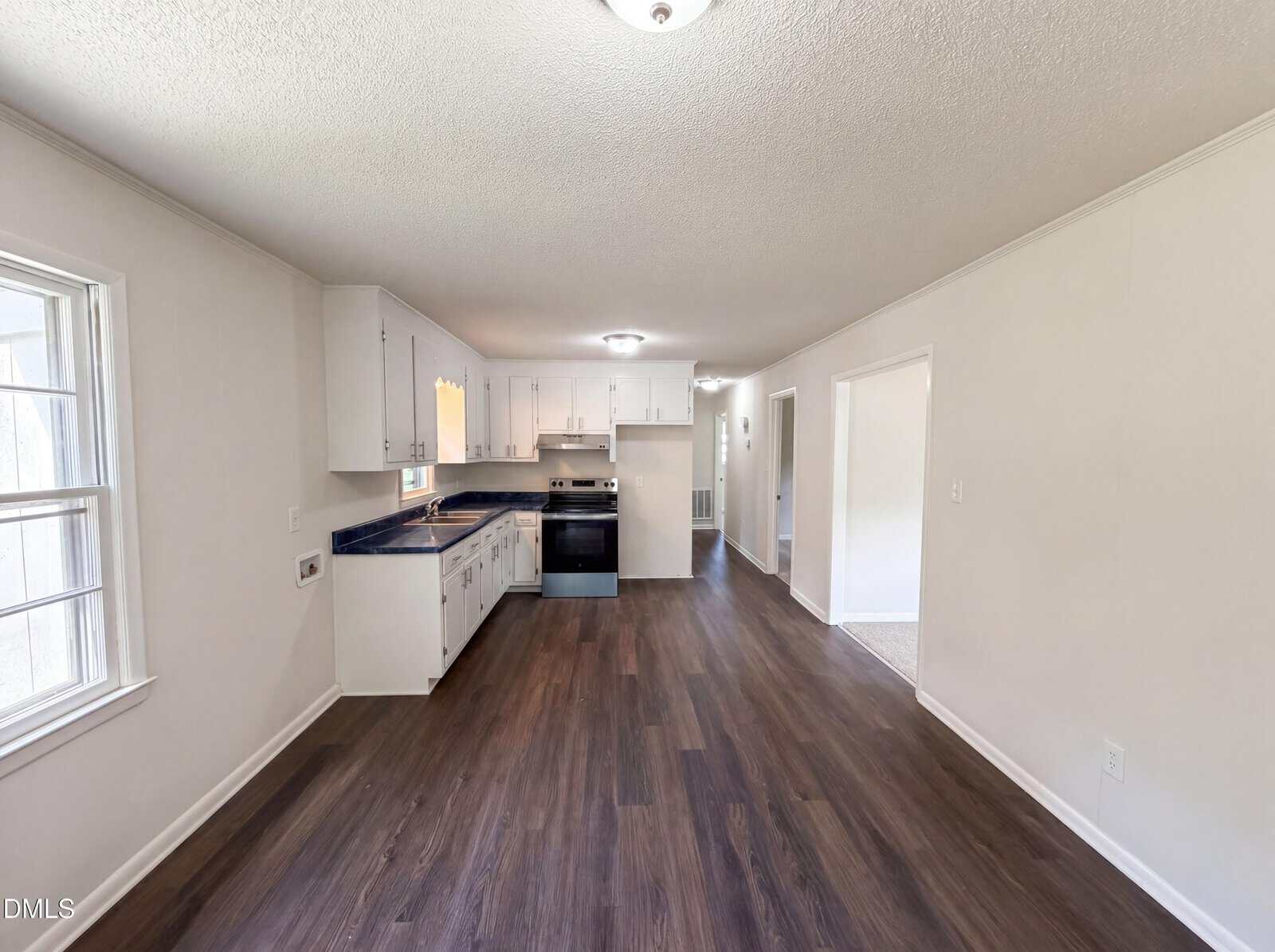Bright kitchen and dining area with white cabinets, dark countertops, and stainless steel appliances. The space features wood-style flooring, neutral walls, and ample natural light from a large window.