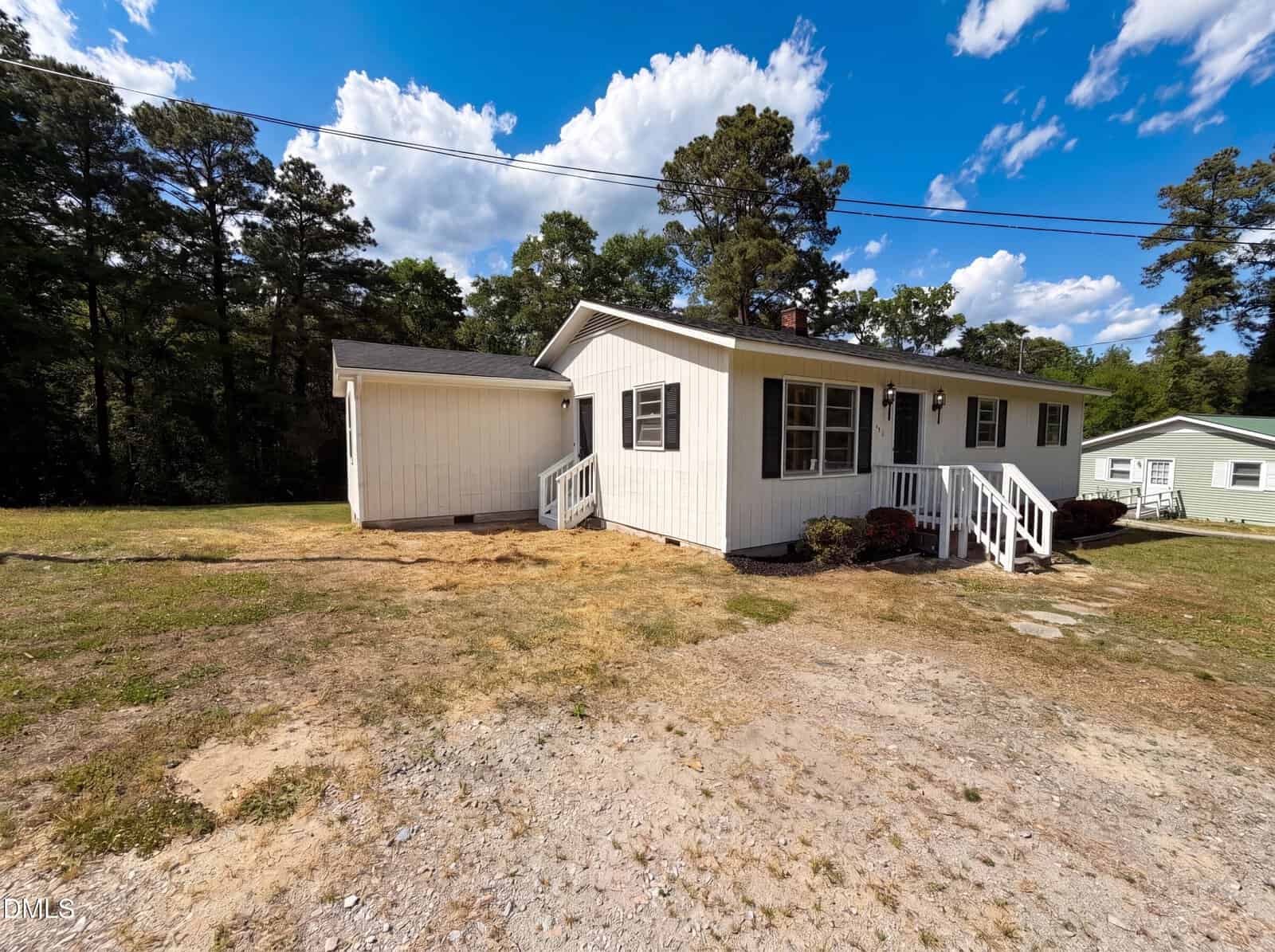 A white, single-story house with dark shutters and two front porches sits on a patchy grass and gravel yard, surrounded by tall trees under a blue sky with scattered clouds.