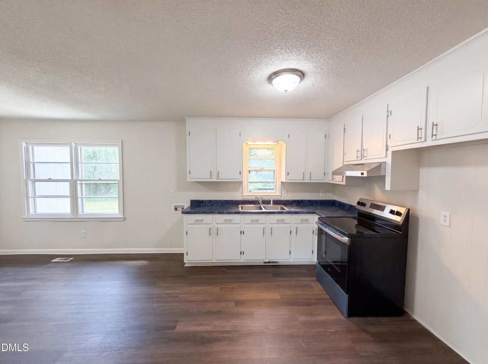 A bright kitchen with white cabinets, a black stove, stainless steel sink, and dark countertops. There are two windows, one above the sink and another on the adjacent wall, letting in natural light. The flooring is dark wood.