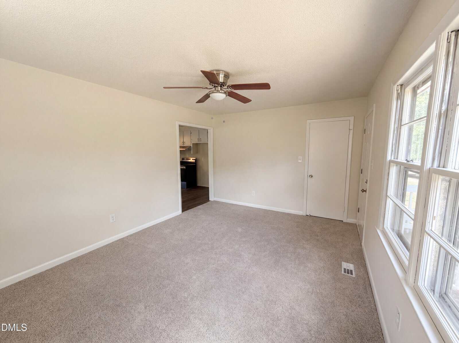 A bright, empty room with beige carpet, white walls, a ceiling fan, three windows, and a door. An open doorway reveals a kitchen with dark flooring and cabinets in the background.