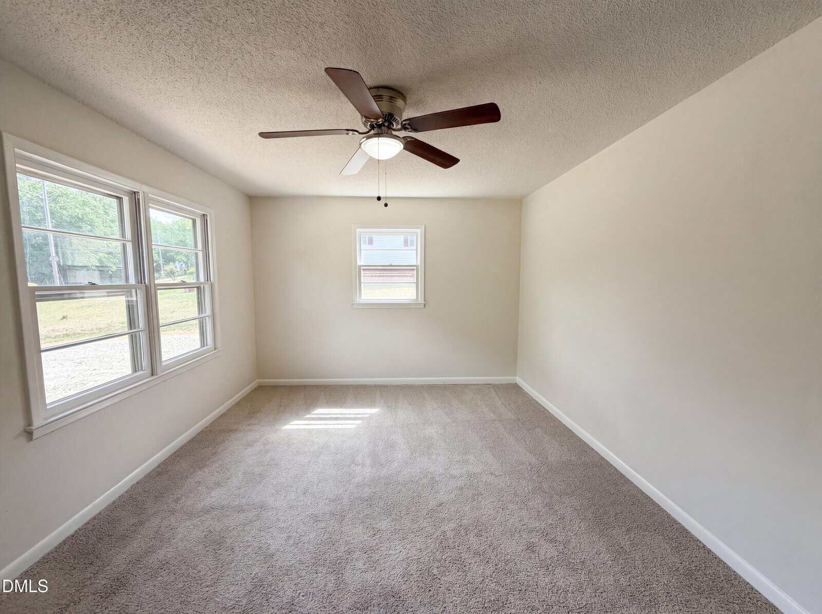 Empty room with beige carpet, white walls, a ceiling fan with light, and three windows letting in natural light. The room appears clean and bright.