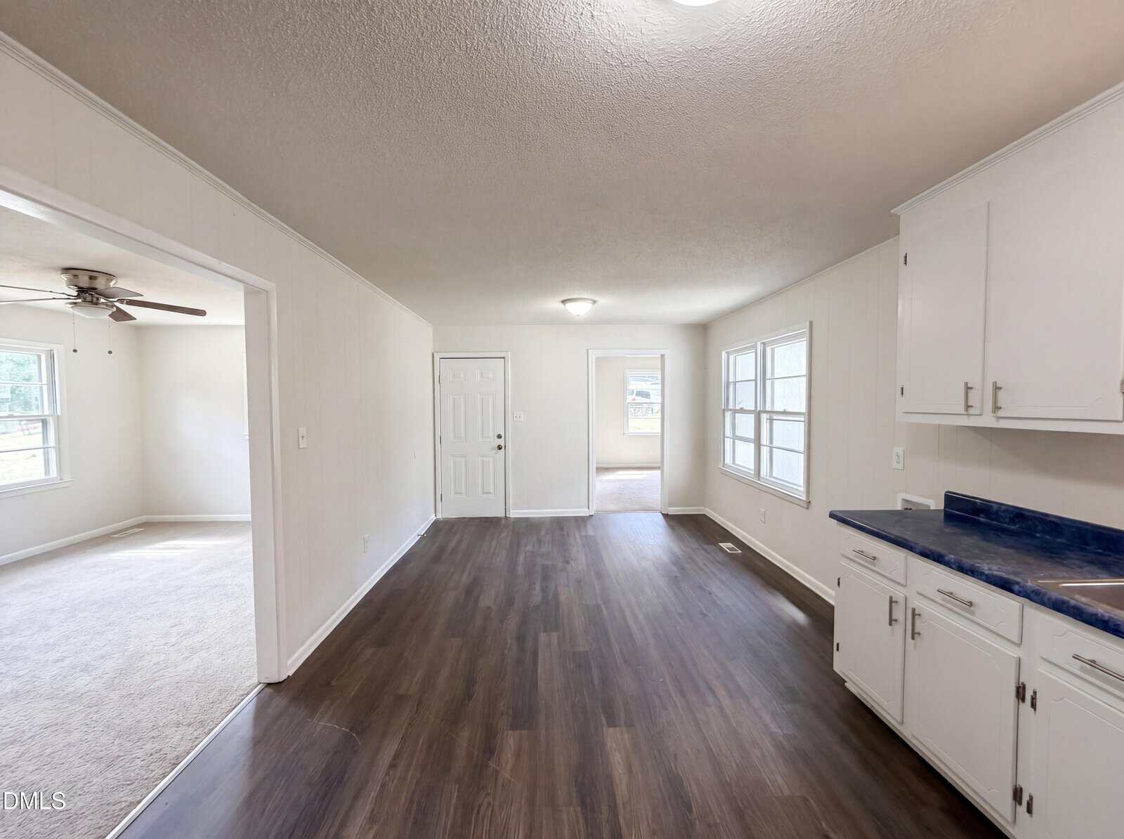 A bright, empty room with dark wood floors, white walls, and white kitchen cabinets. Large windows let in natural light, and a door is visible at the far end. An adjacent carpeted room has a ceiling fan.
