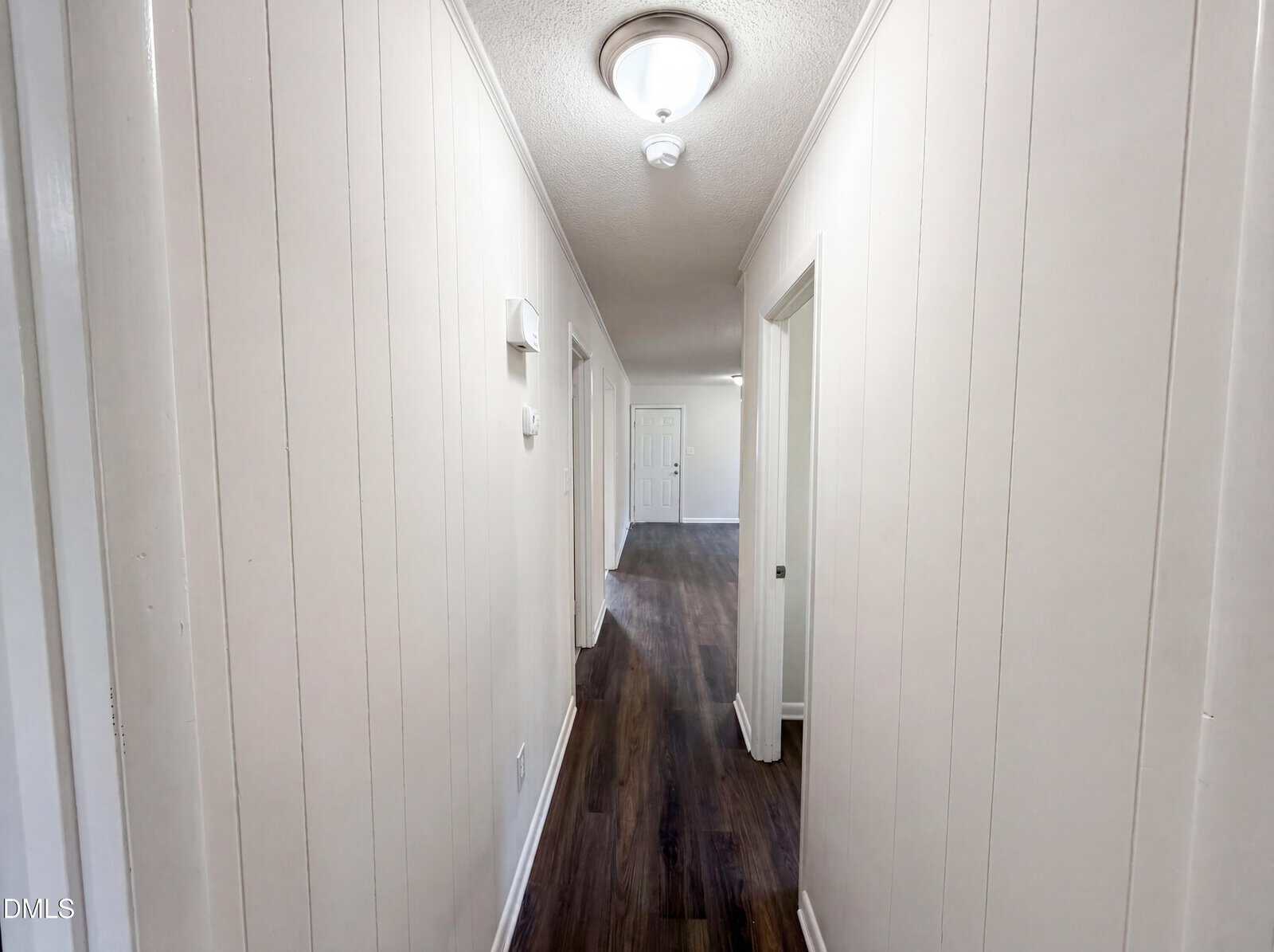 A long hallway with white paneled walls, dark wood flooring, a ceiling light, and several doorways leading to different rooms; a closed white door is visible at the end.