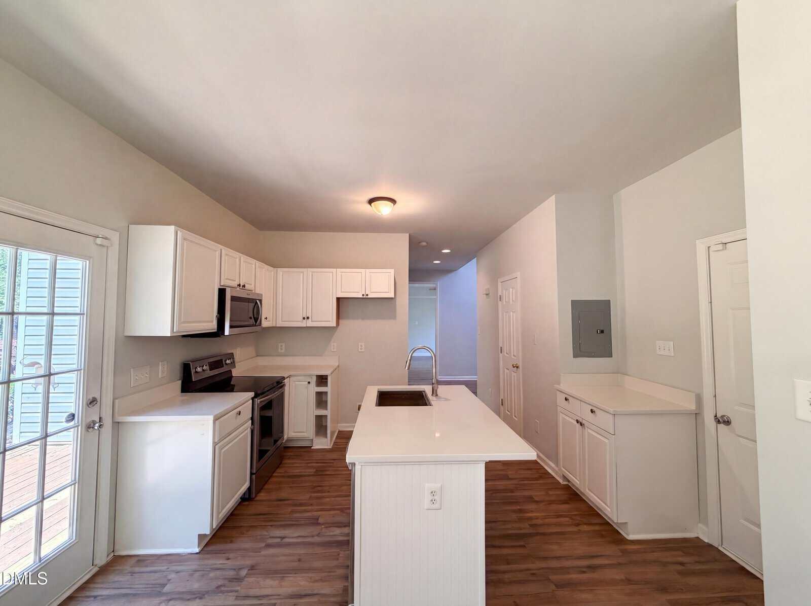 Bright, modern kitchen with white cabinets, stainless steel appliances, a central island with sink, wood-style flooring, and a door with glass panels leading outside. Neutral walls and ceiling lights complete the space.