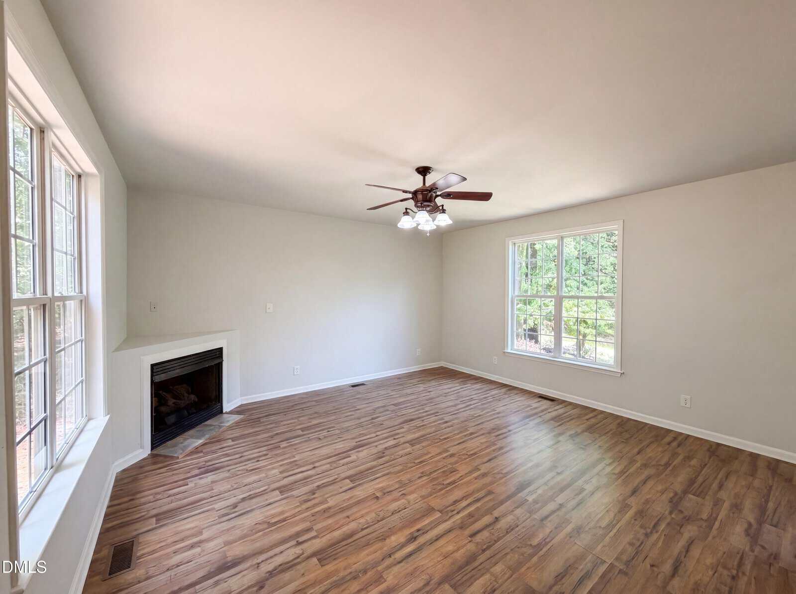 Bright, empty living room with wood flooring, large windows, a ceiling fan with lights, and a corner fireplace. White walls and natural light create an open, airy atmosphere.