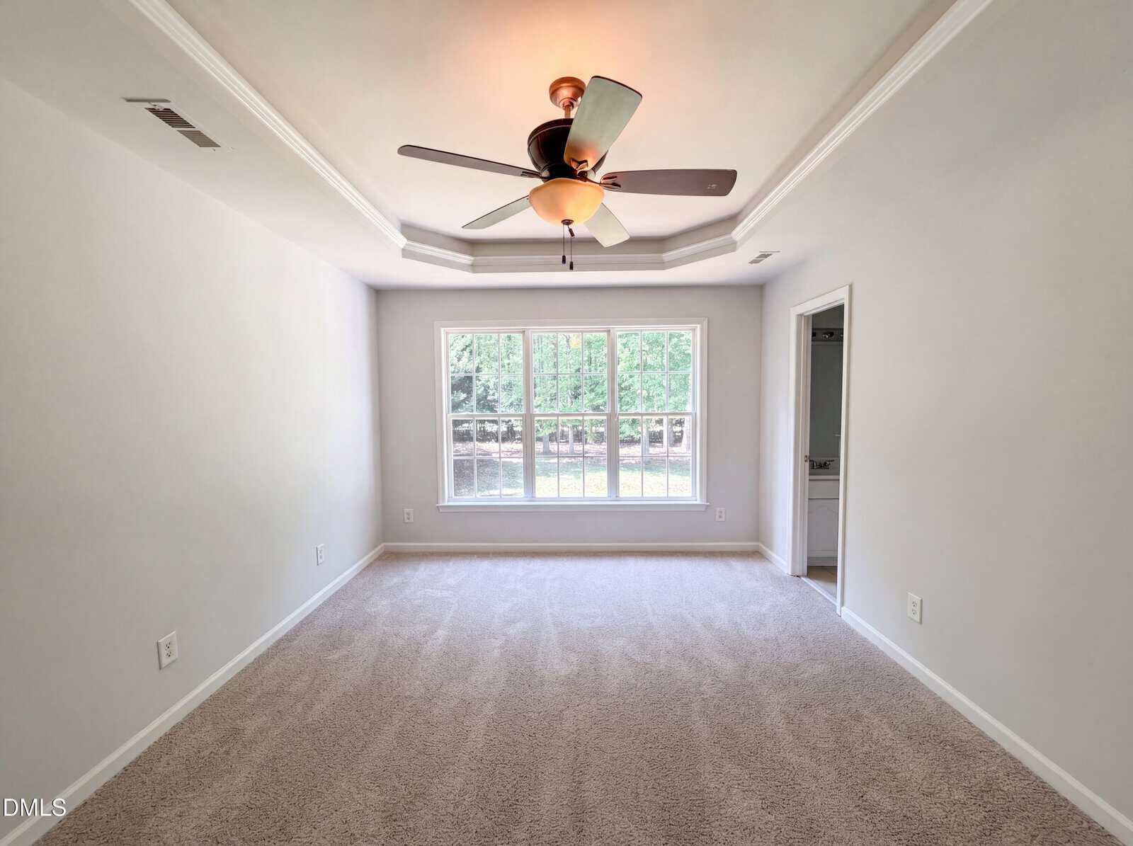 A spacious, empty room with light carpet, white walls, a ceiling fan with lights, and a large window overlooking a green yard. There is a doorway leading to another room on the right.