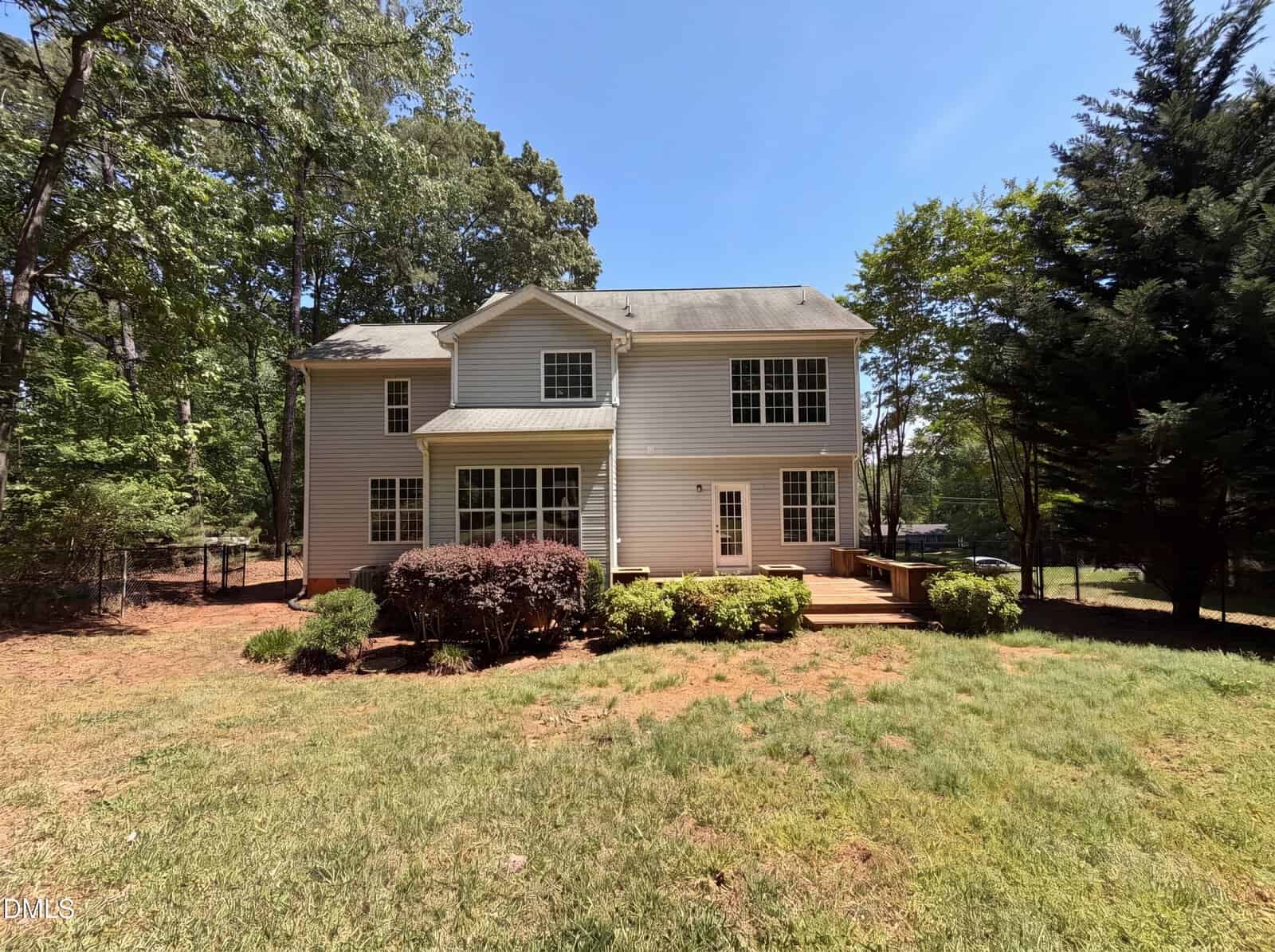 A two-story beige house with white trim and several large windows, surrounded by trees and bushes, with a wooden deck and a spacious, sunlit backyard.