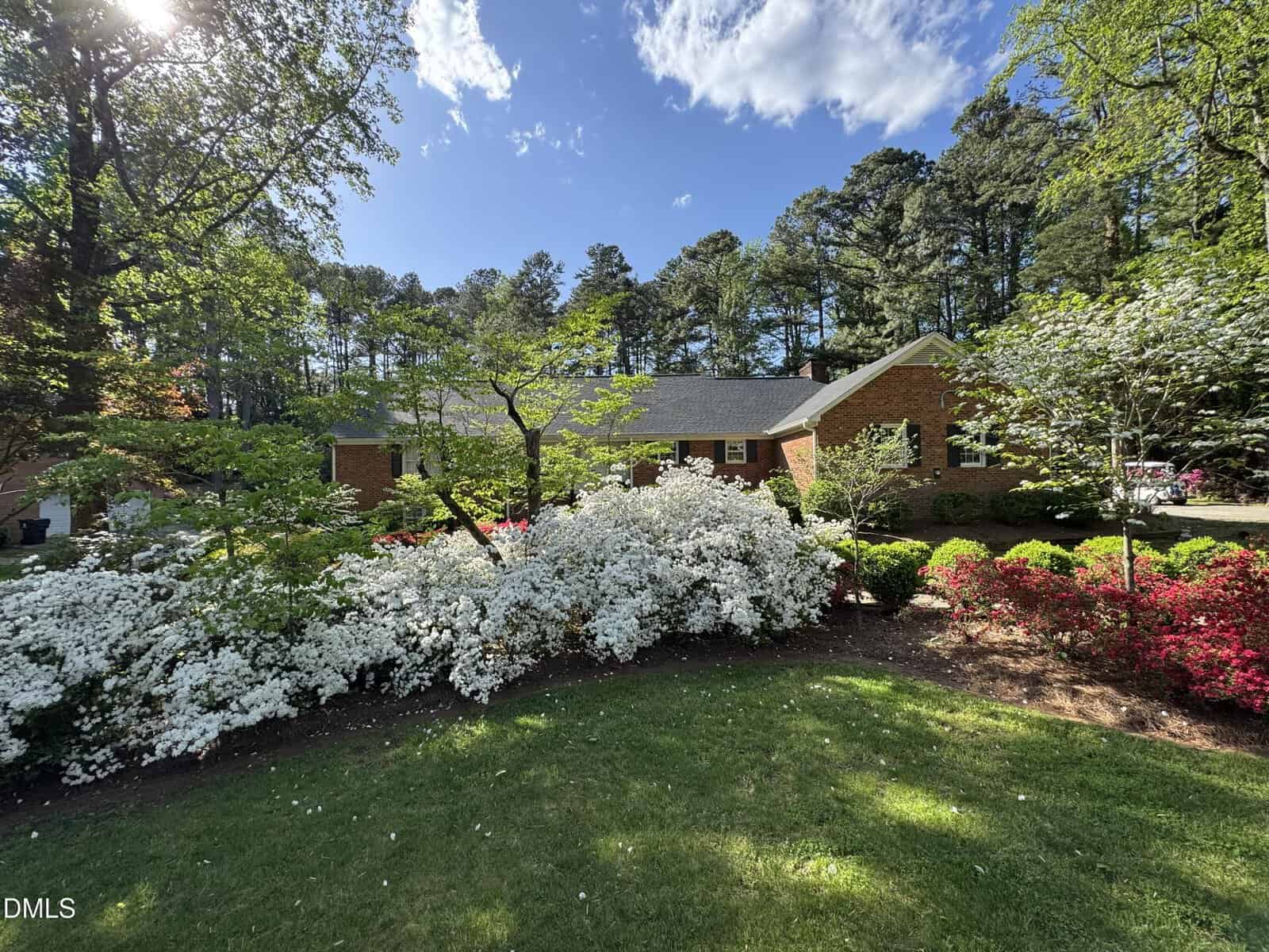 Single-story brick house surrounded by lush green trees, vibrant white and red flowering bushes, and a well-kept lawn under a bright blue sky with scattered clouds.