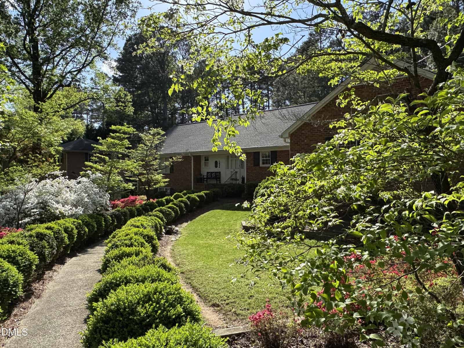 A brick house with a covered front porch is surrounded by lush greenery, manicured bushes, blooming flowers, and a curved pathway leading to the entrance under tall trees on a sunny day.