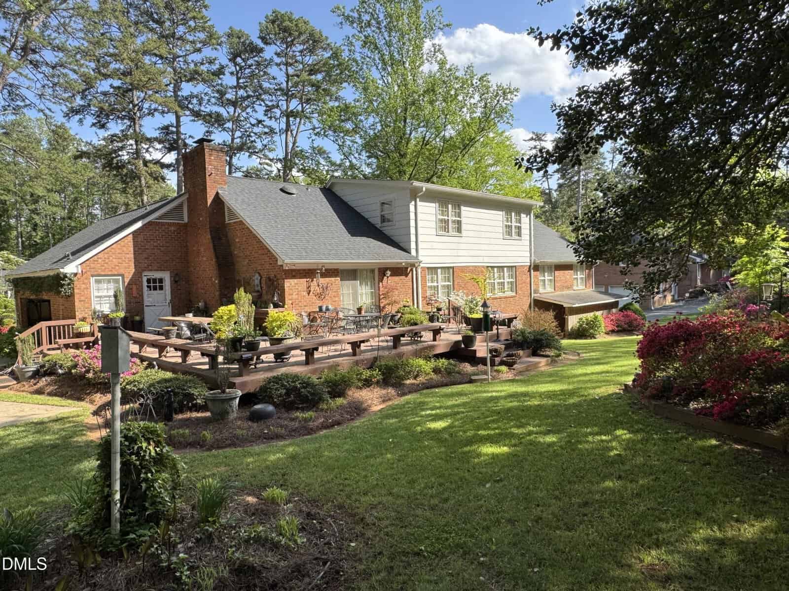 A large two-story brick house with a wooden deck, surrounded by landscaped gardens, flowering bushes, and tall trees under a partly cloudy sky.